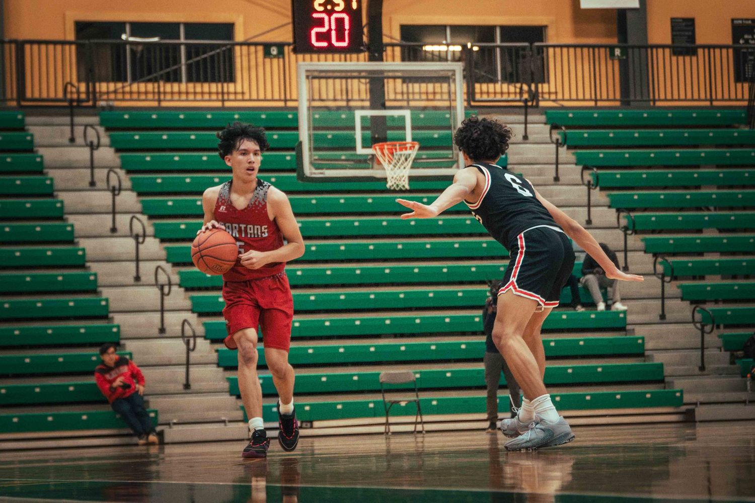 Basketball player in red uniform faces defender in black during game with green stadium seating.