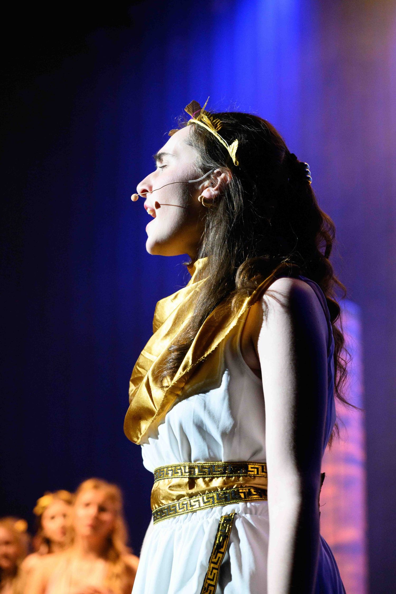 Performer in white Greek-style dress with gold belt performs under blue stage lighting.