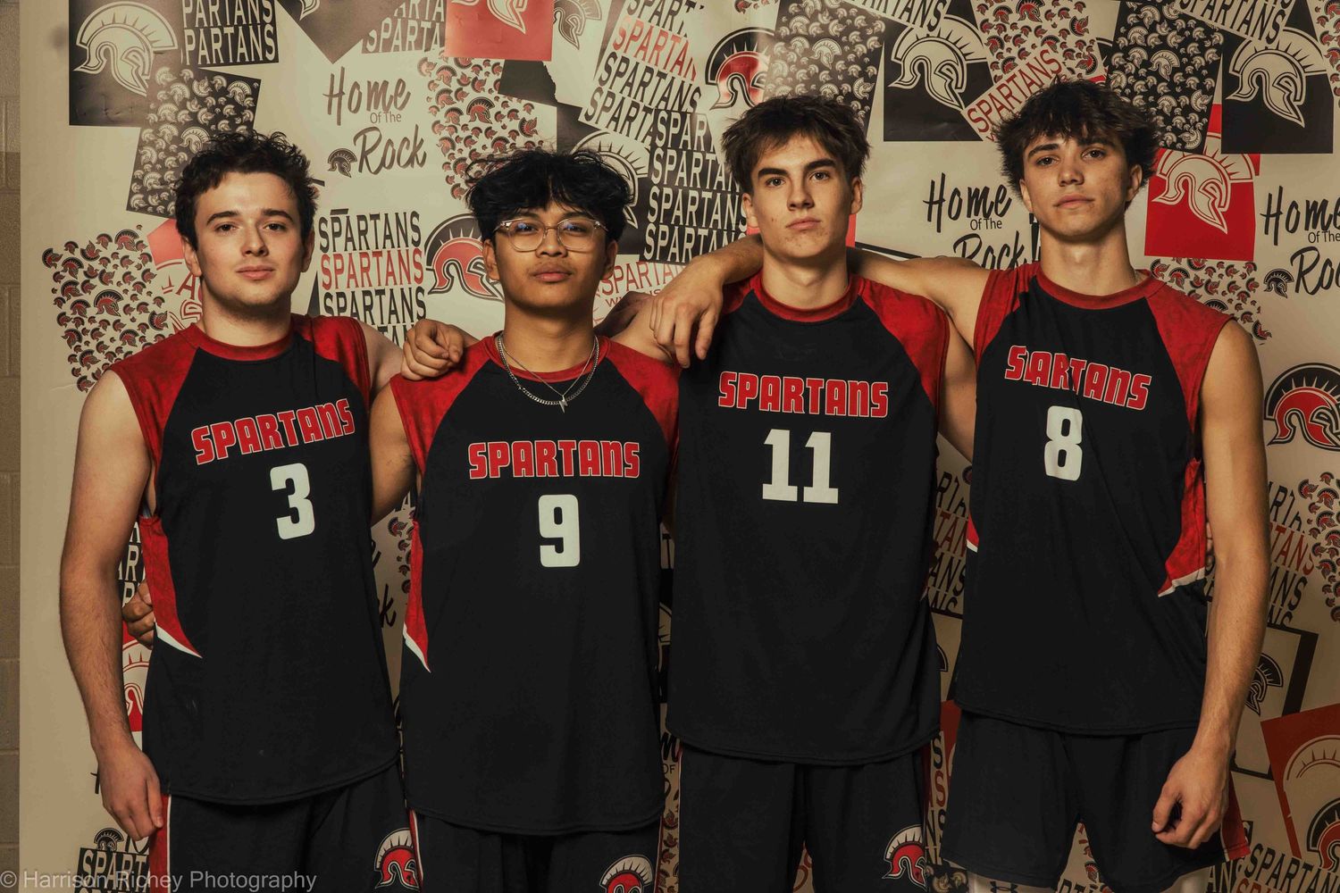 High school volleyball team wearing black uniforms poses together against a decorative wall background.