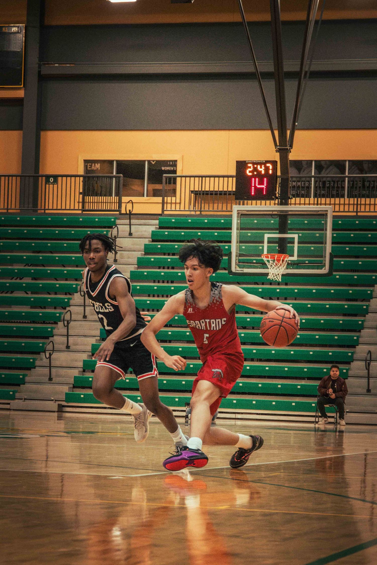 Players engage in basketball game action on court with stadium scoreboard visible.