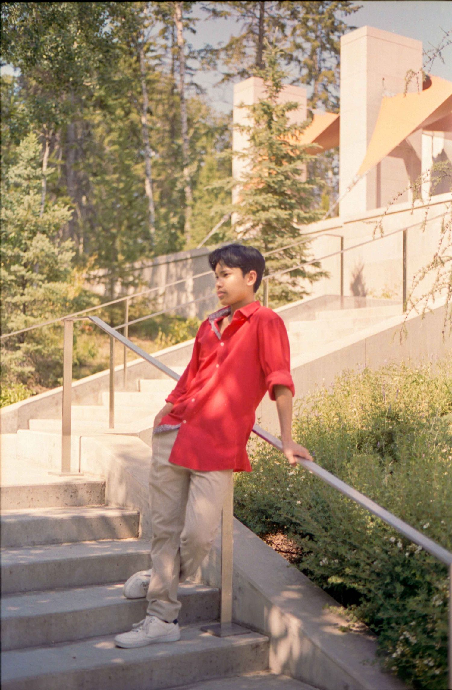 Person wearing bright red shirt and khakis poses on outdoor concrete steps near greenery.