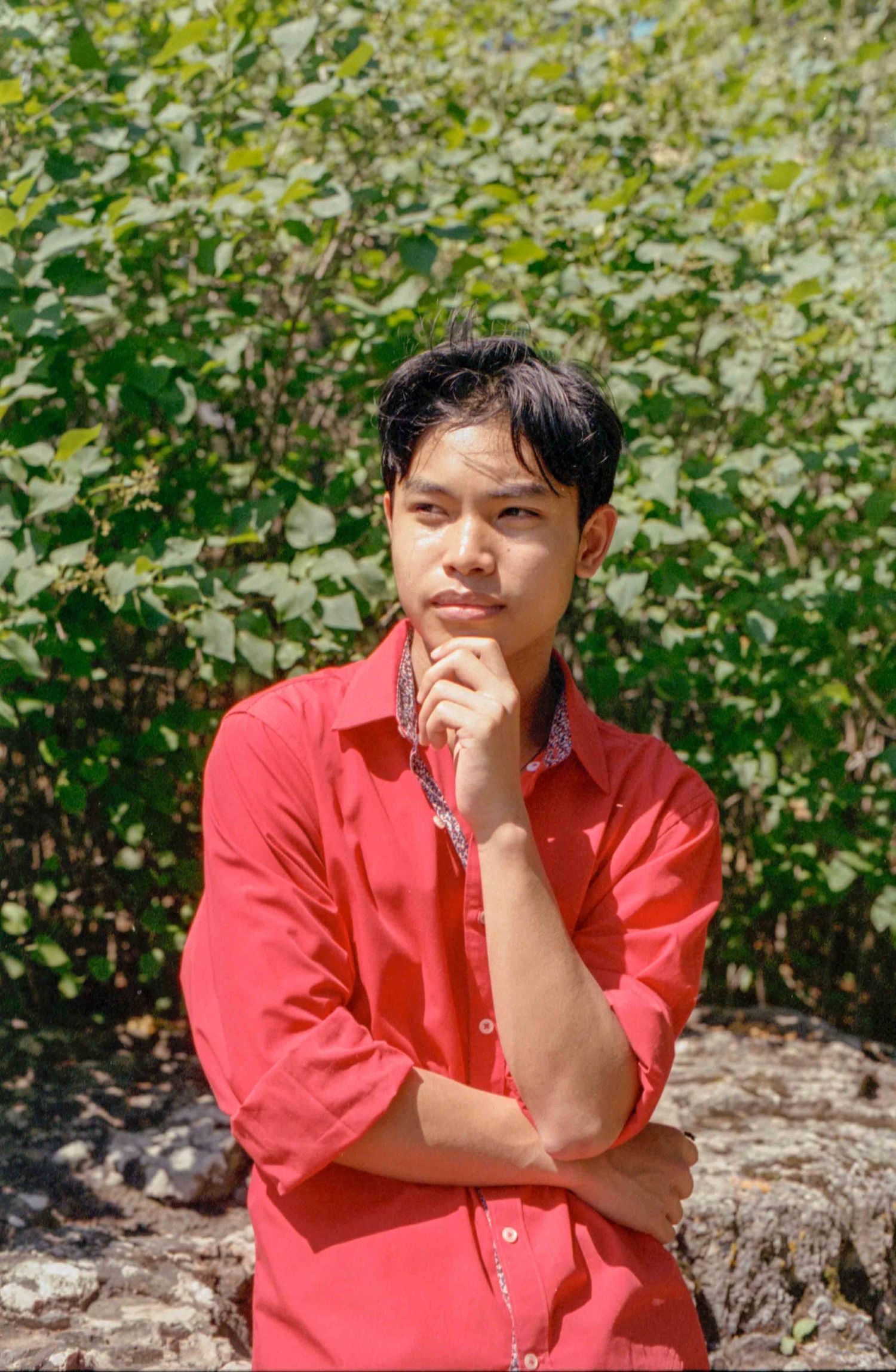 Person in coral button-down shirt poses thoughtfully against green foliage background.