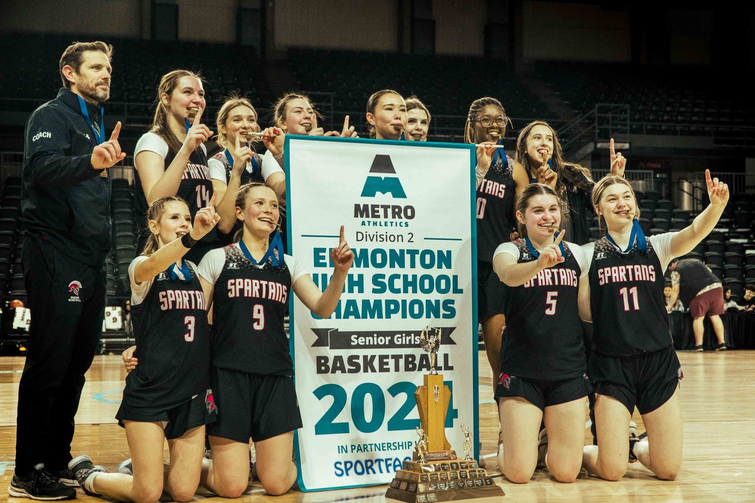 Basketball team celebrates with their 2021 Metro championship banner in a dark arena.