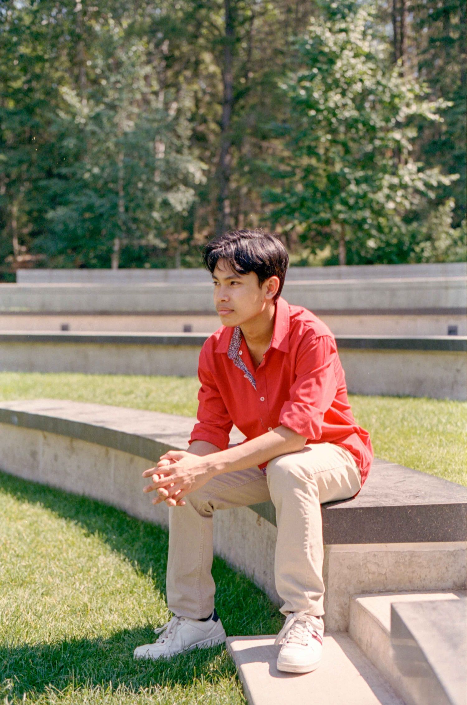 Young person in red shirt and khaki pants sits casually on concrete steps in a garden setting.