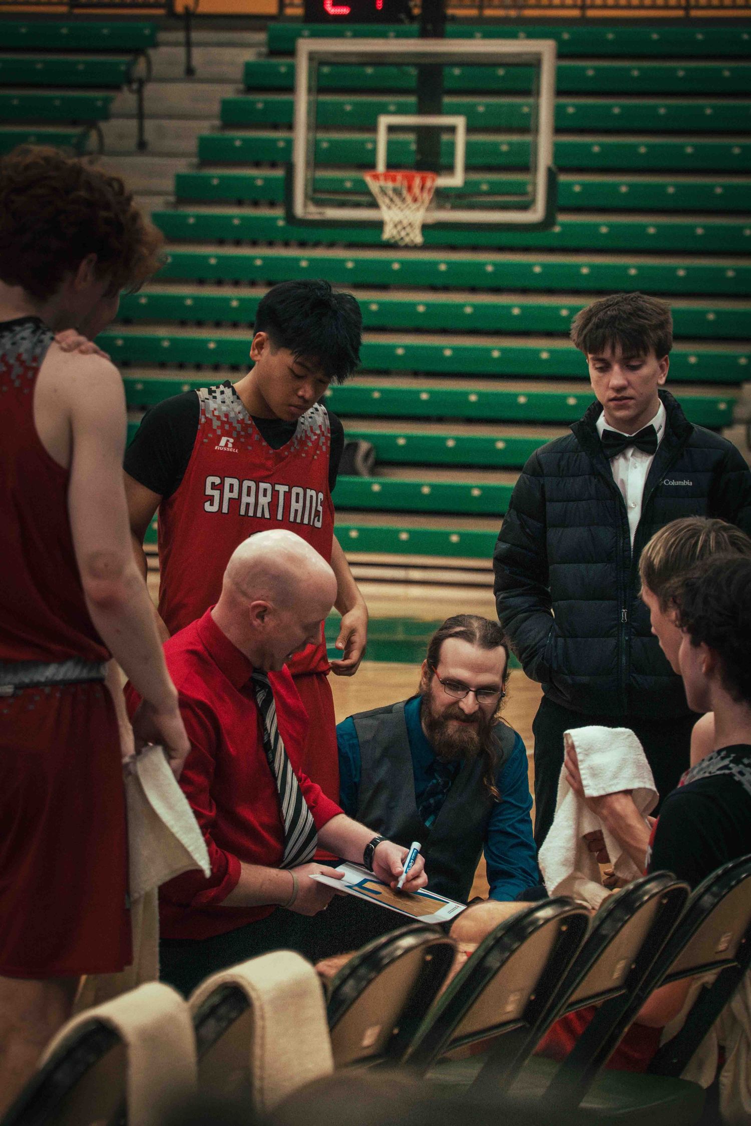 Basketball team huddles around coach reviewing strategy during timeout break.