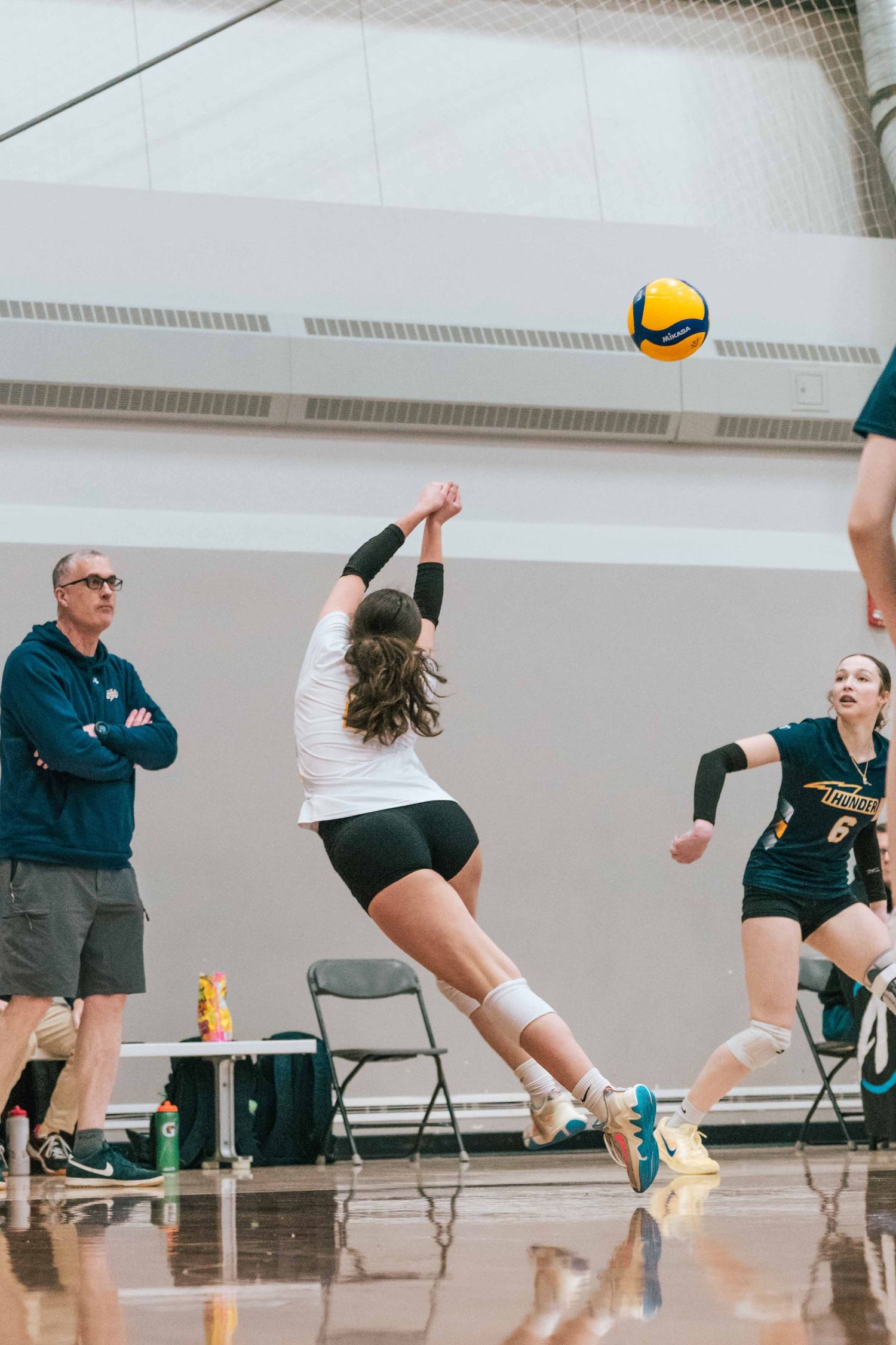 Volleyball player jumps to hit ball during match while coach observes from sideline.