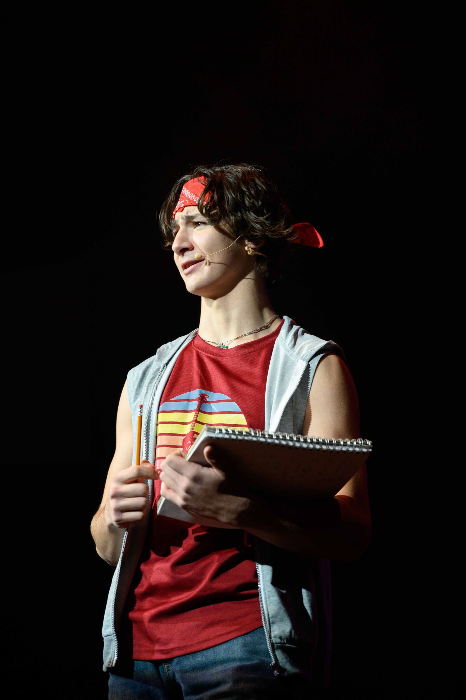A student in a red shirt and vest takes notes while studying in dramatic lighting.
