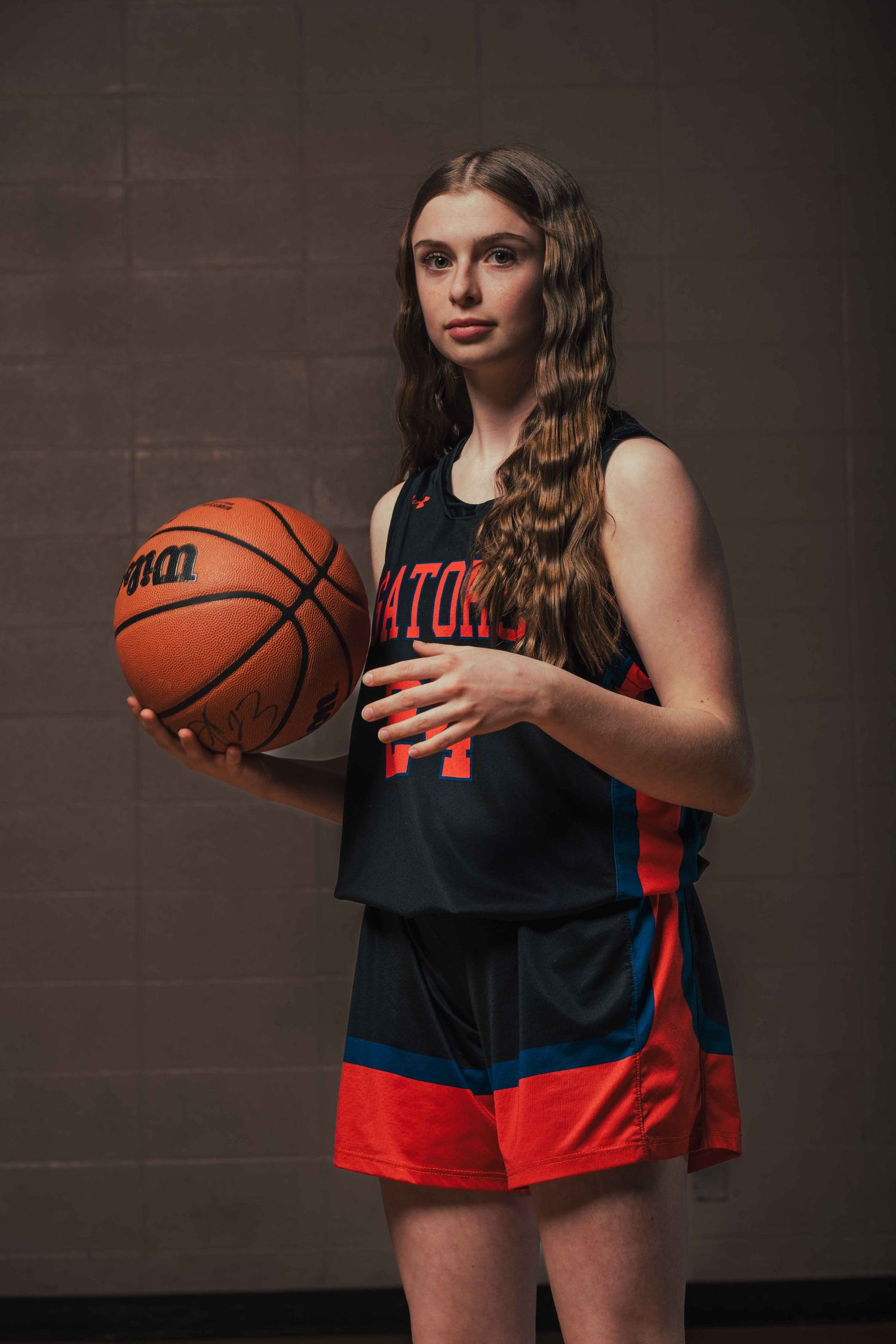 Basketball player in black and red uniform poses with ball against dark background.