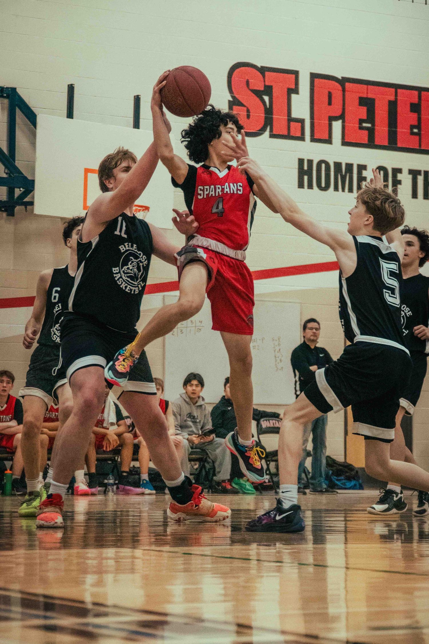 Youth basketball players in red and black uniforms jump and compete for rebounds.