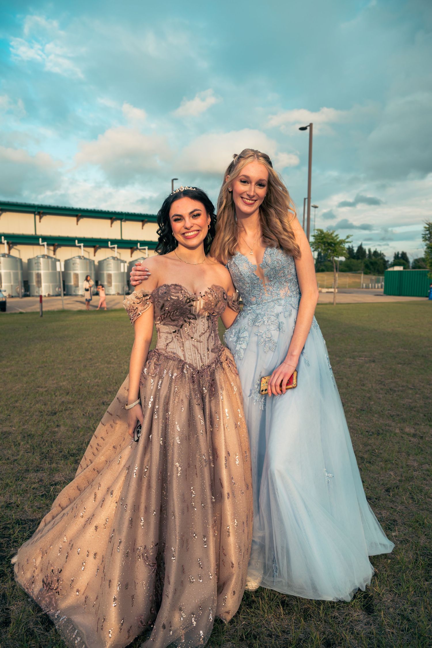 Two people in formal dresses, one in sparkly bronze and one in light blue, posing on a grassy field at dusk.