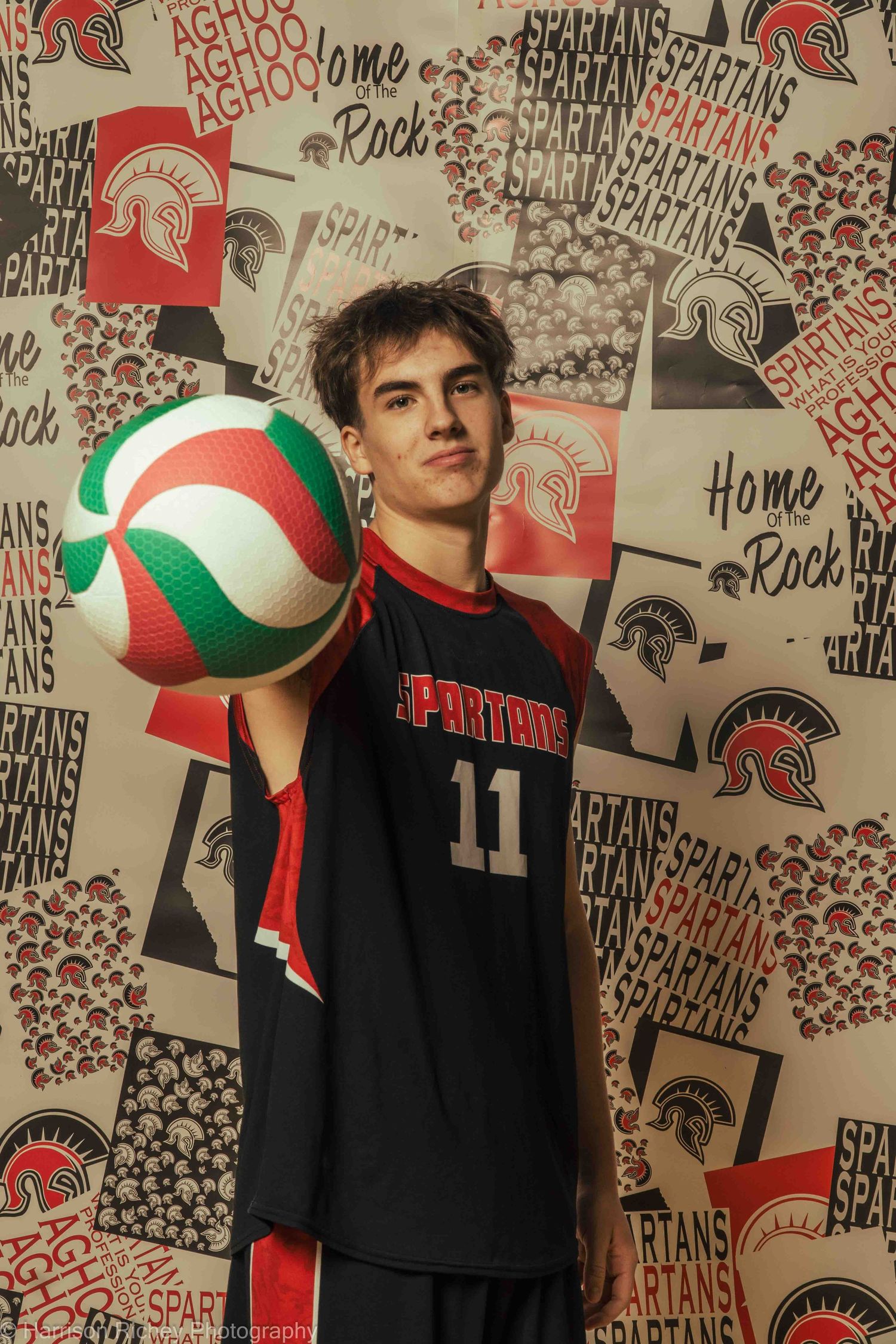 Basketball players holding volleyballs pose in black uniforms against team-themed backdrop.