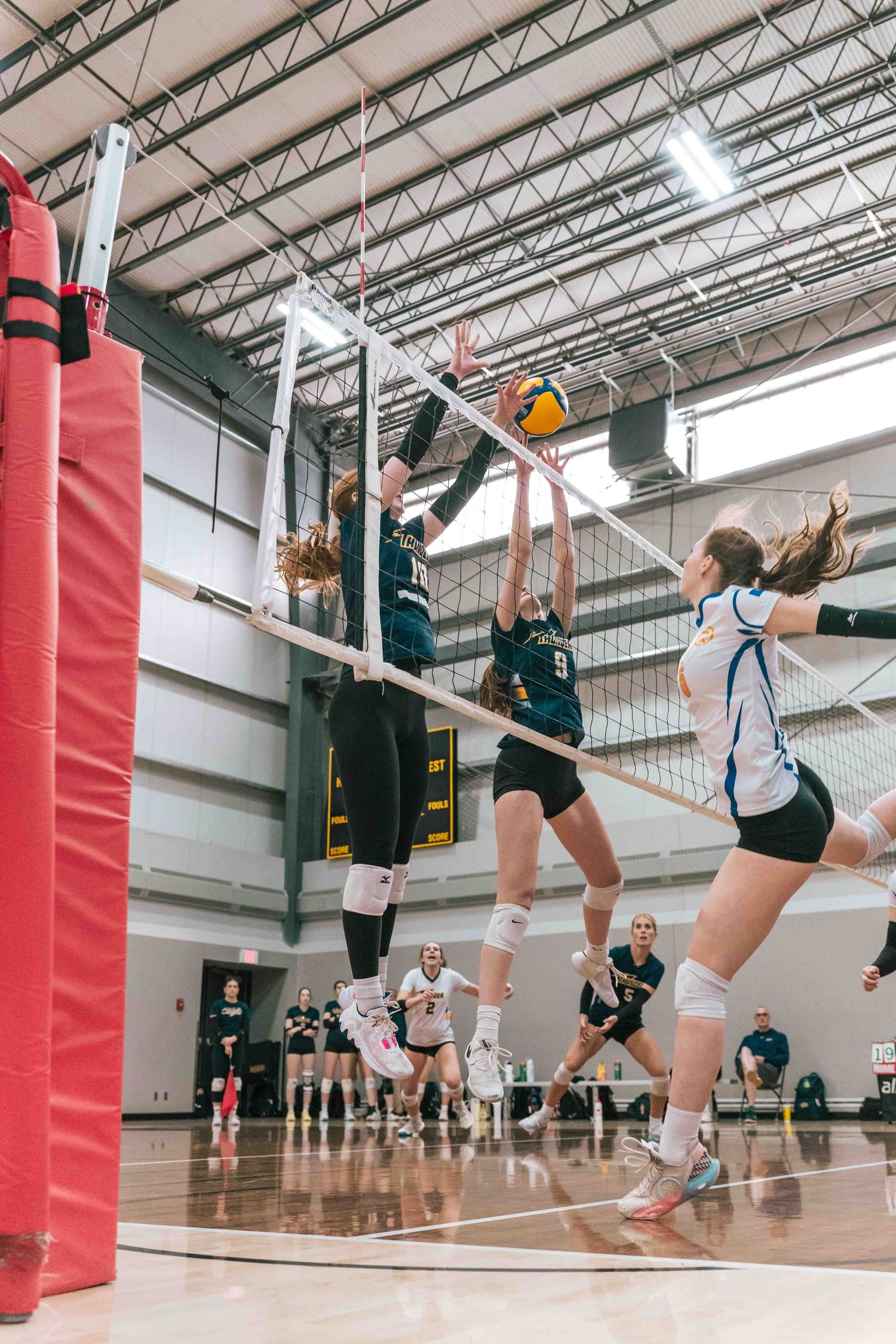 Indoor volleyball players leap high at the net for a block during an intense match.