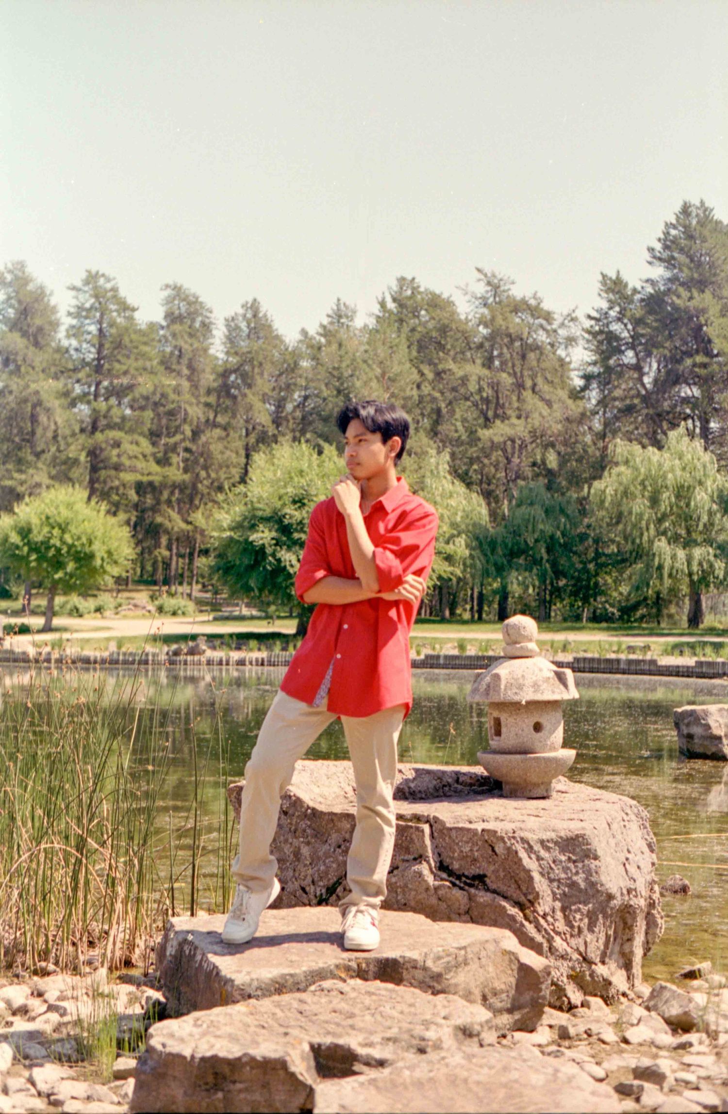 Person in red outfit stands on decorative rocks near a tranquil pond with trees in background.