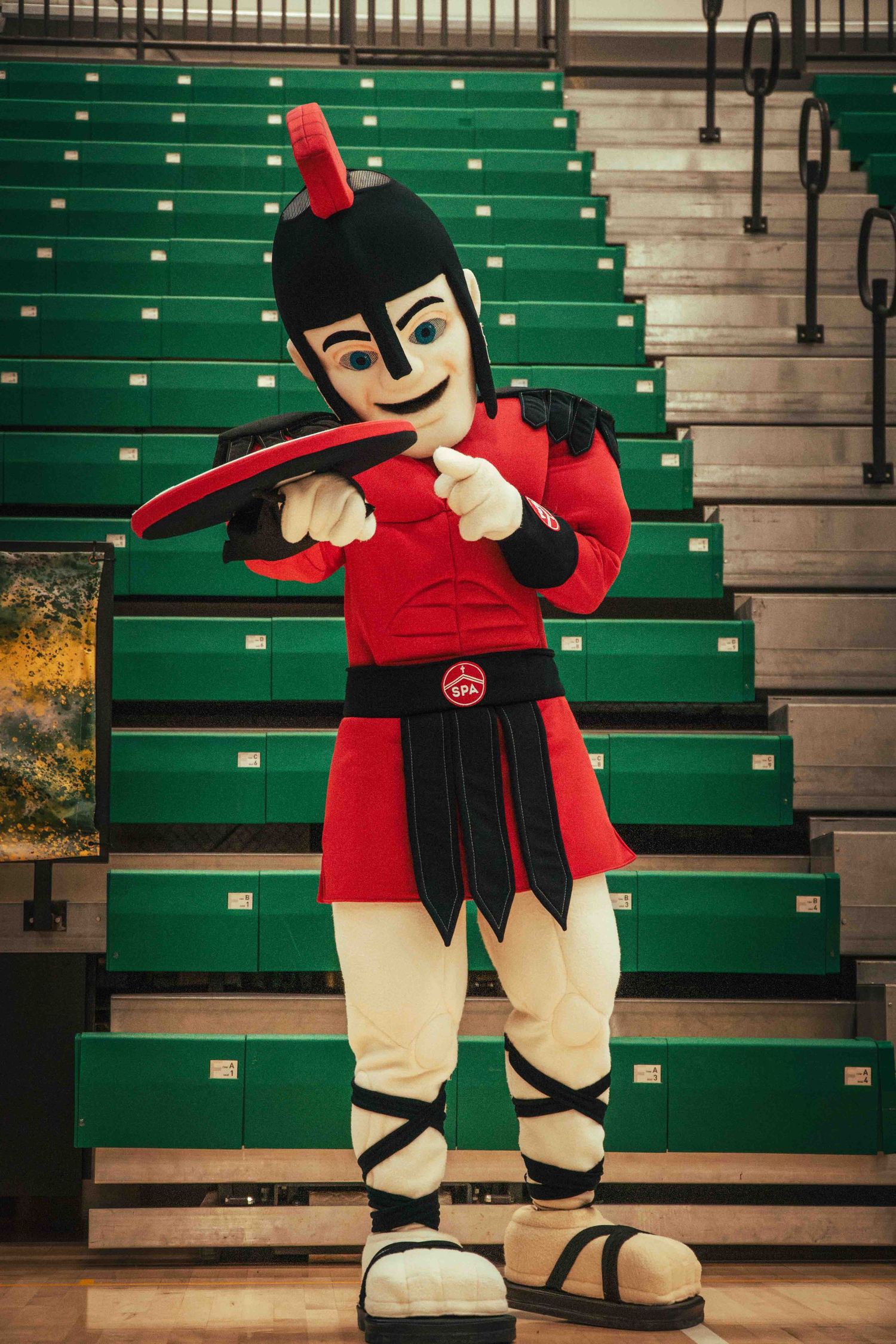 School mascot in red and black warrior costume poses in front of green bleachers.