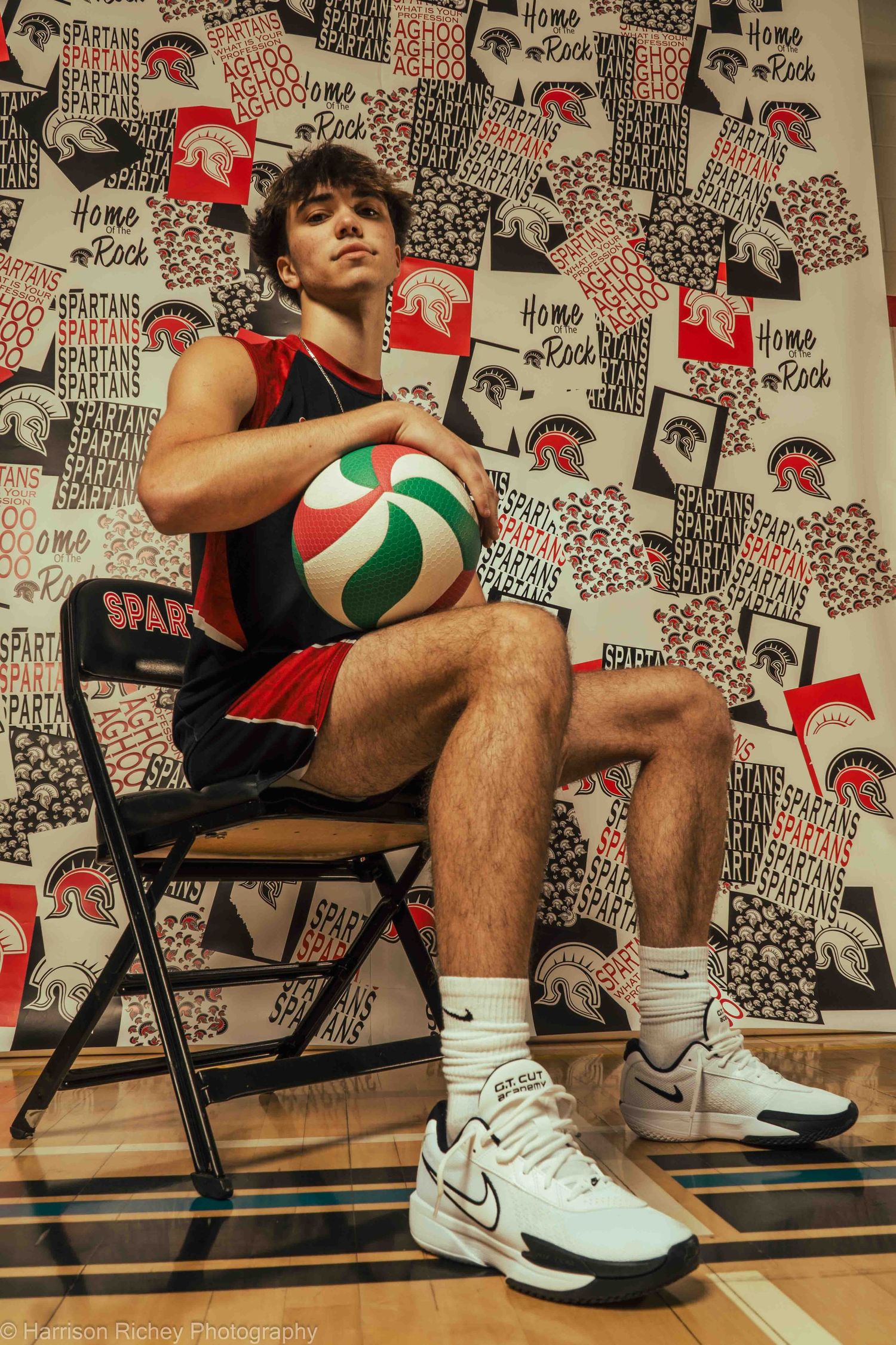Basketball players in team uniforms sit on chairs against patterned wall holding volleyballs.