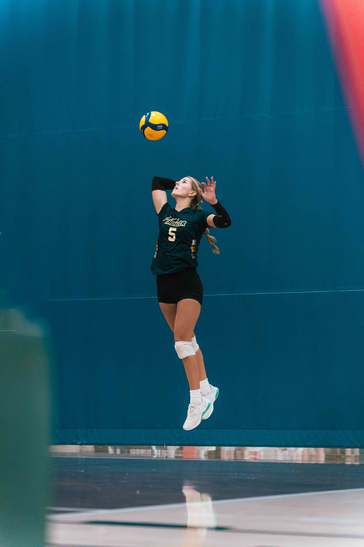 Player in dark uniform leaps to serve volleyball during indoor match.