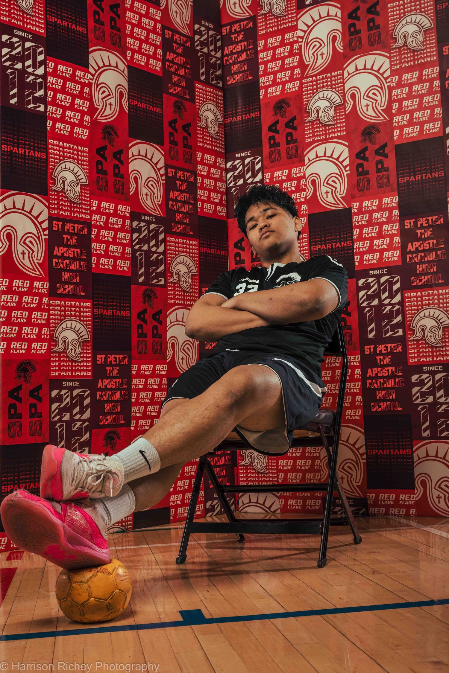 Athlete relaxes in chair with handball at feet against red patterned wall with team logos.