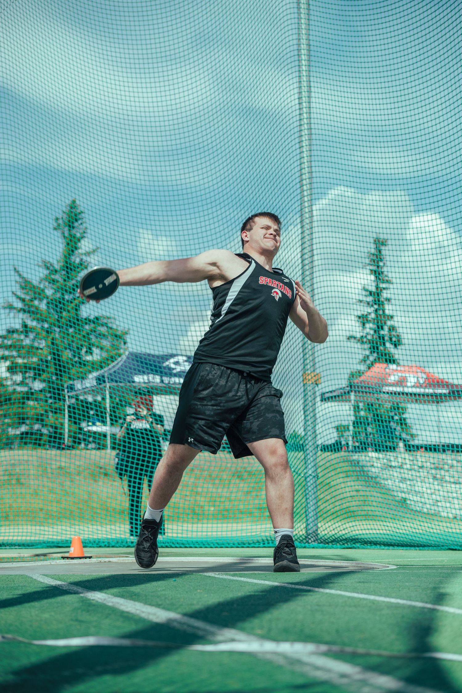 Athlete performing a discus throw sequence in an outdoor stadium with protective netting and evergreen trees in background.