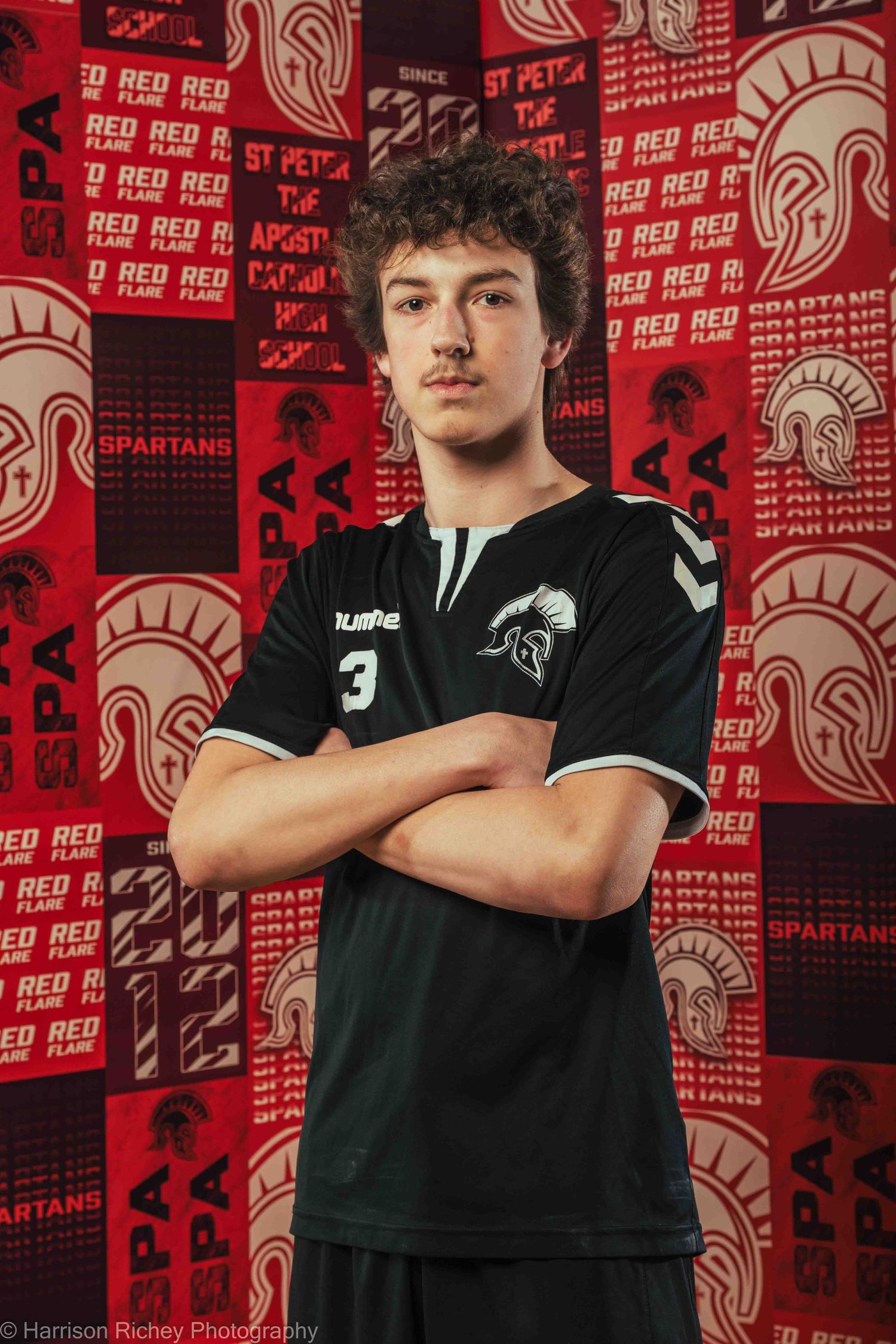 Handball player in black uniform poses confidently against red team logo backdrop.
