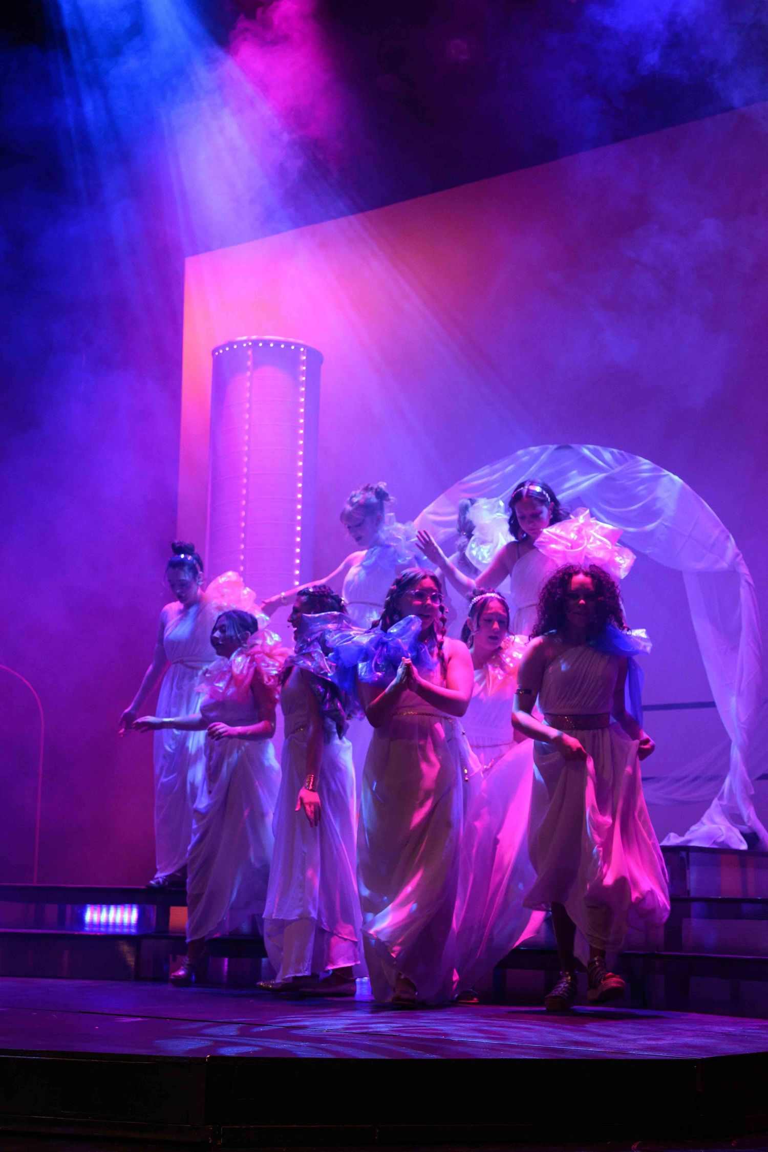 Ethereal dance performance with multiple performers in flowing white costumes under blue and purple lighting.