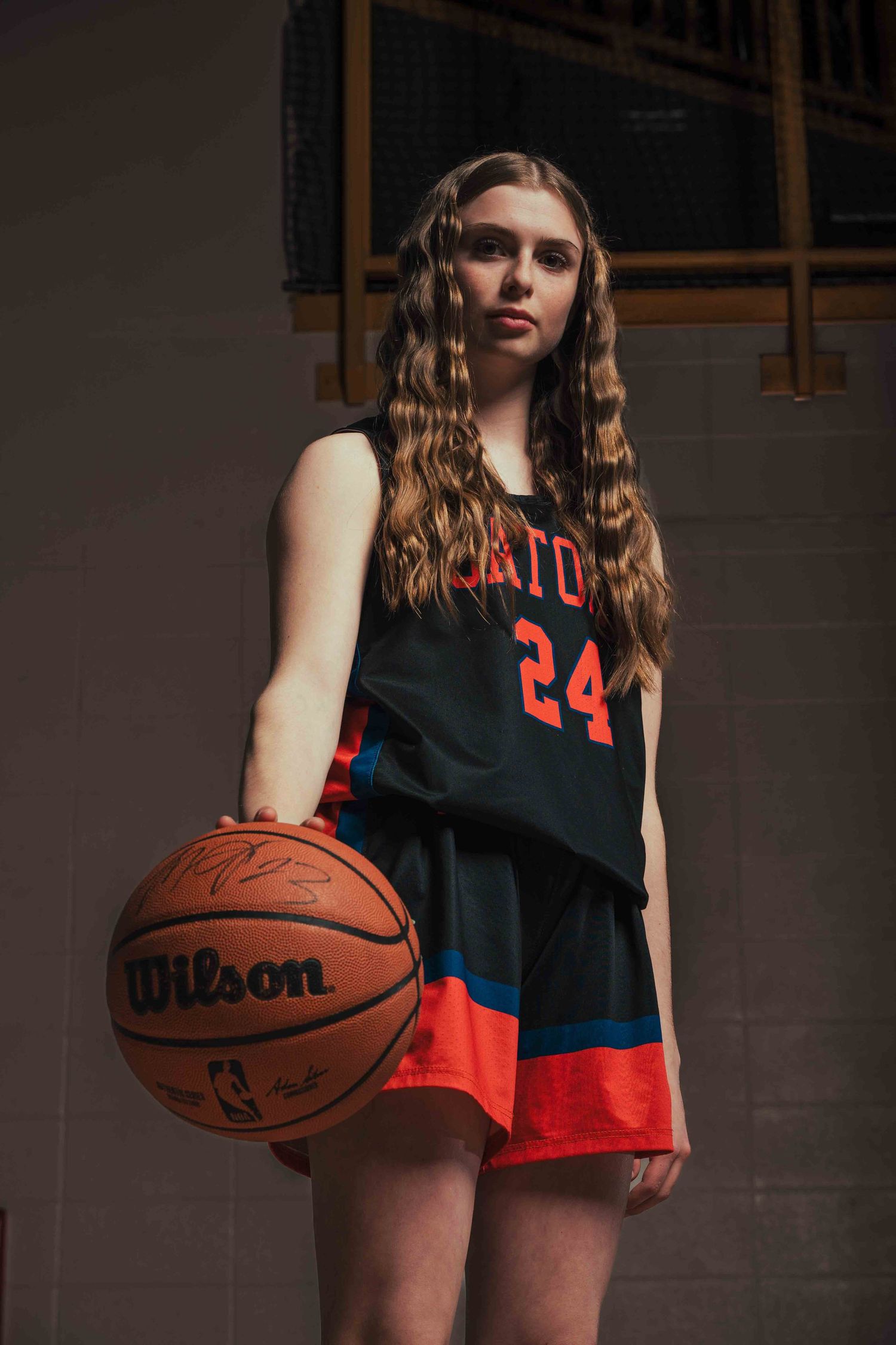 Basketball player in dark jersey holds Wilson basketball in moody lighting.