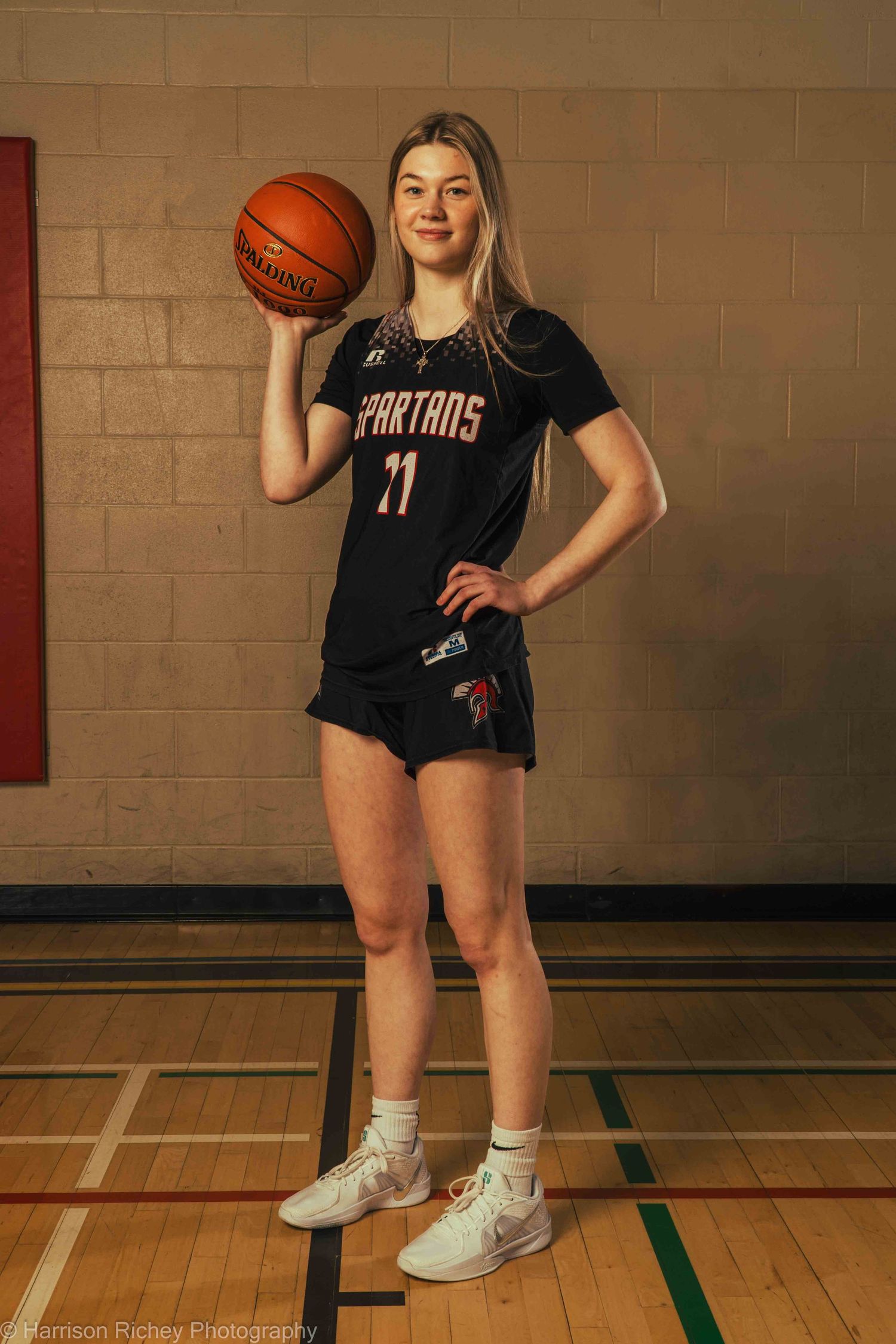 Basketball player in Spartans uniform poses with ball on wooden gym floor.