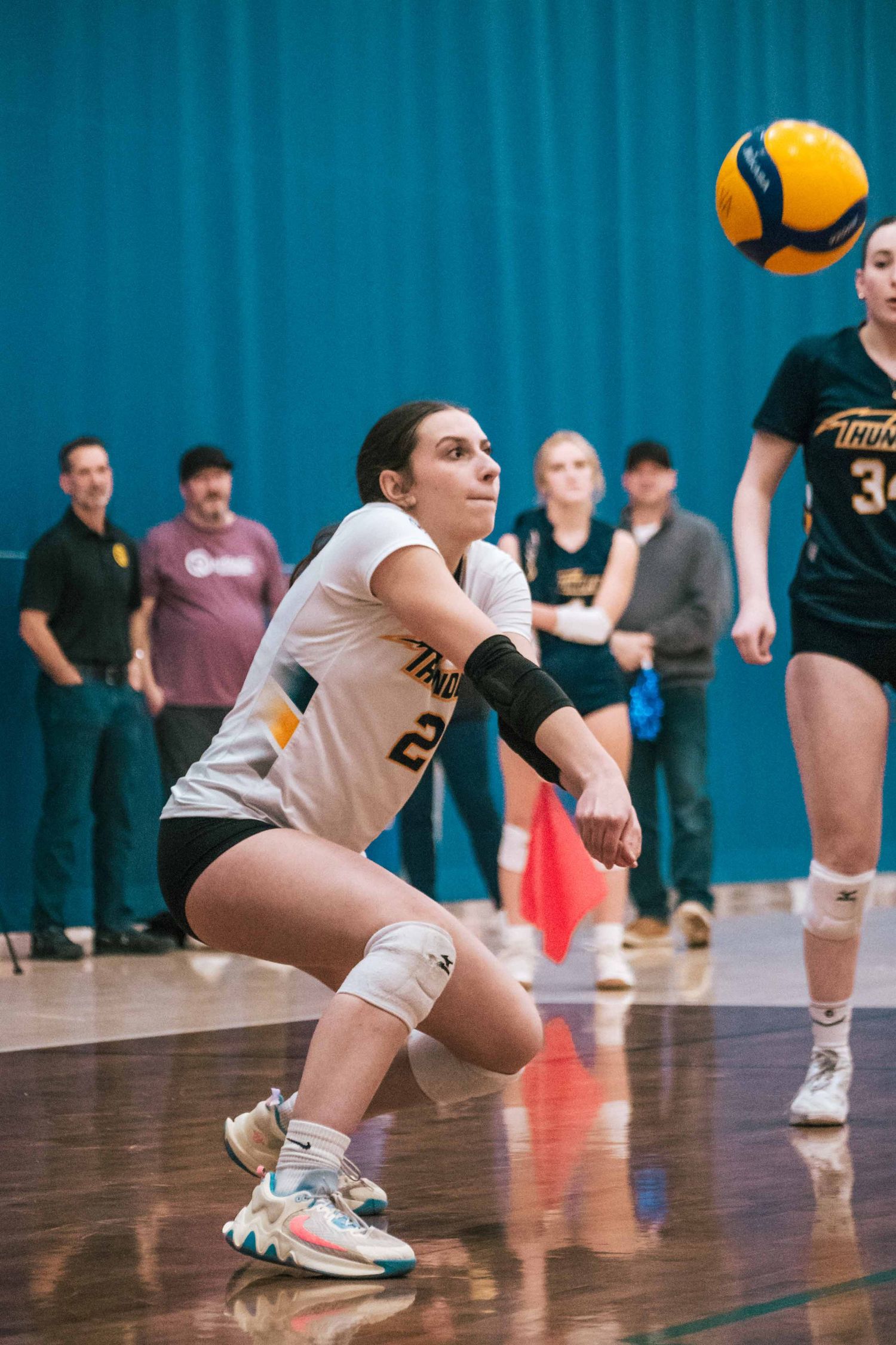 Indoor volleyball player in white jersey prepares to receive a yellow and blue ball on the court.