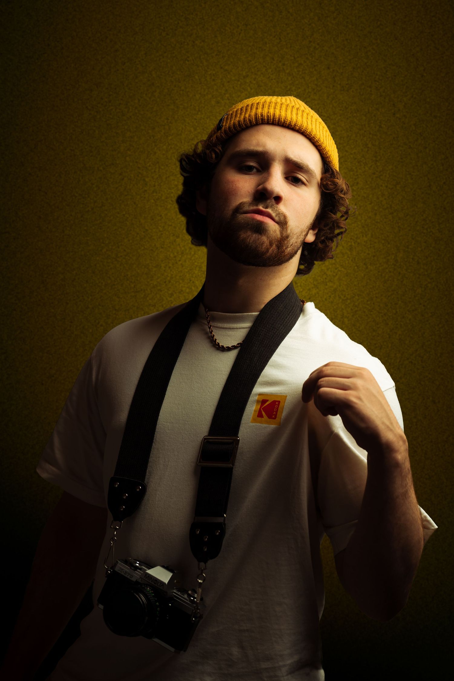 Portrait shot of Harrison Richey in a white t-shirt and yellow beanie against a dark yellow background.