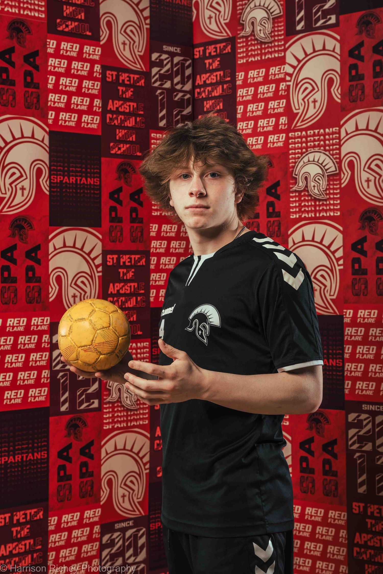Athlete in black uniform holds golden handball against red patterned backdrop with Spartan logos.