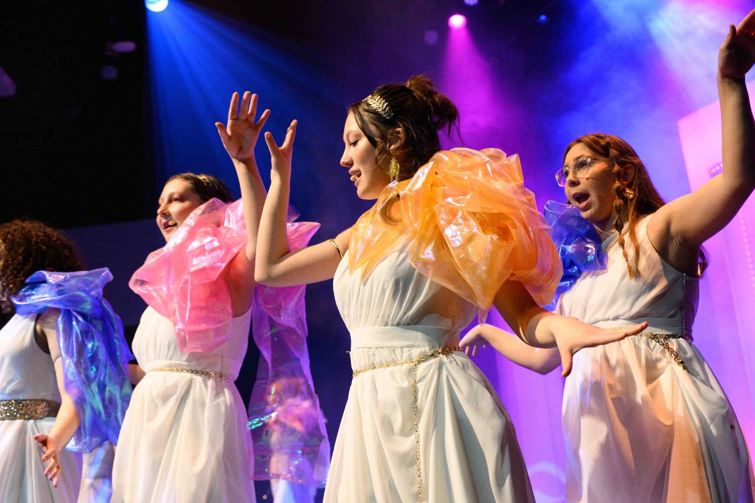 A line of dancers in white dresses perform synchronized movements on stage under colorful lighting.