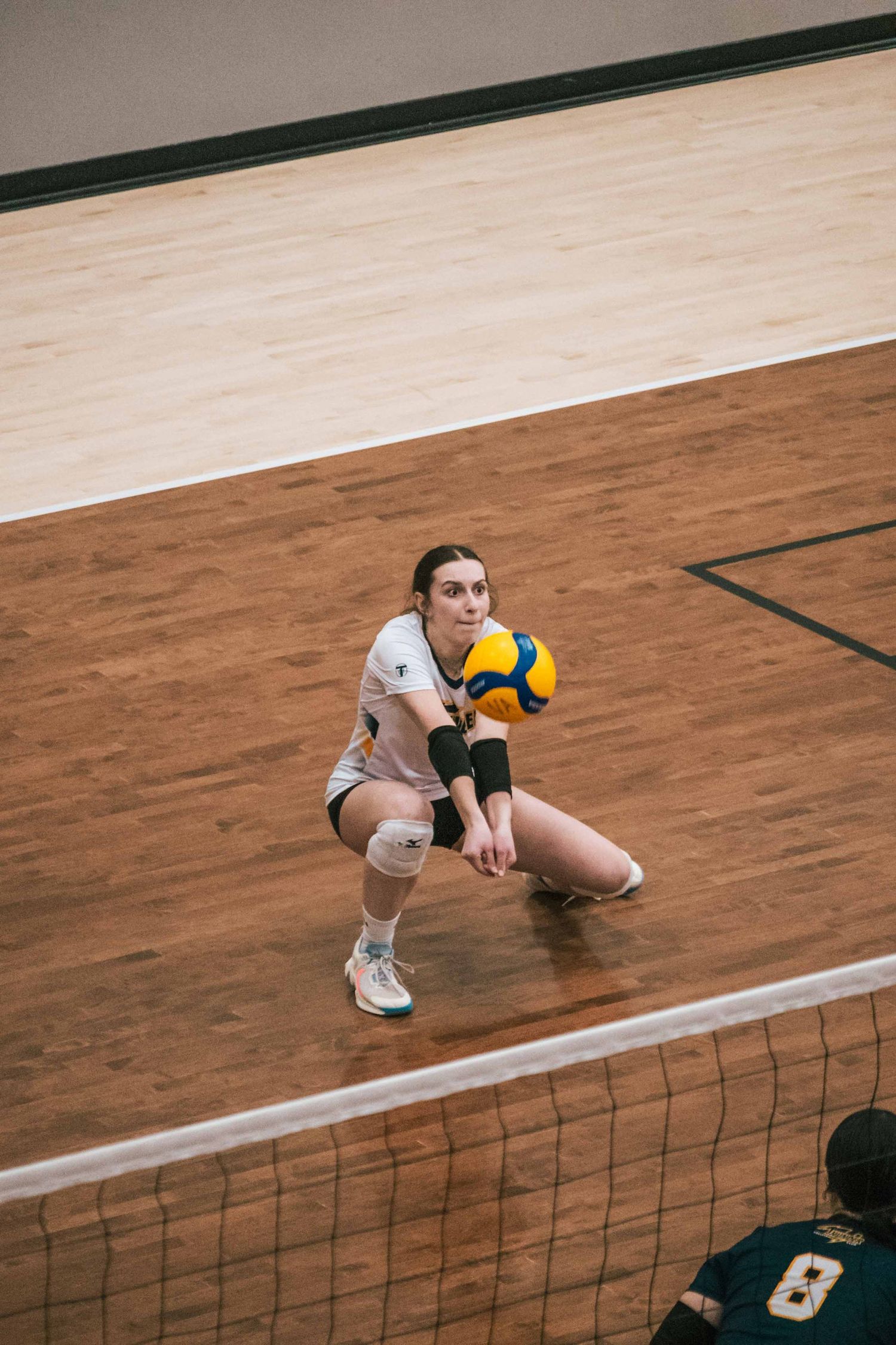 Volleyball player in white uniform crouches low to receive a colorful ball during an indoor match.