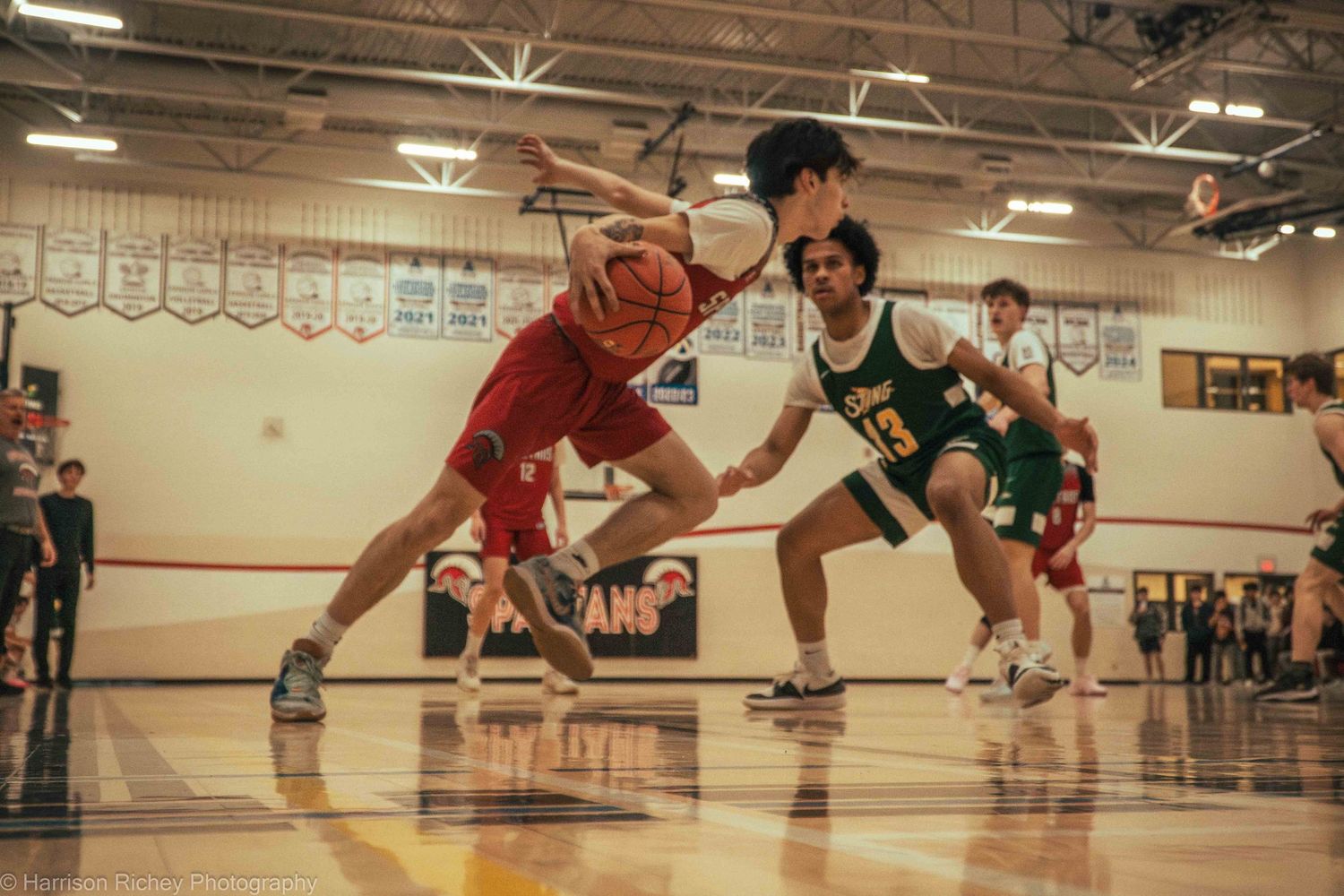 Players in red and black uniforms dive for a loose basketball during an intense game in a gymnasium.
