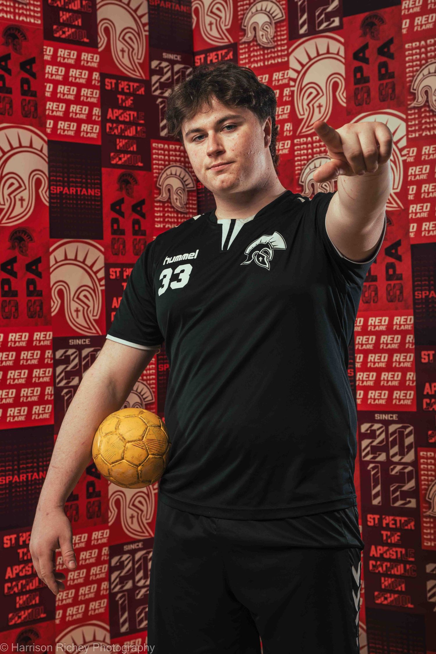 Athlete in black jersey points while holding golden handball against red team banner background.