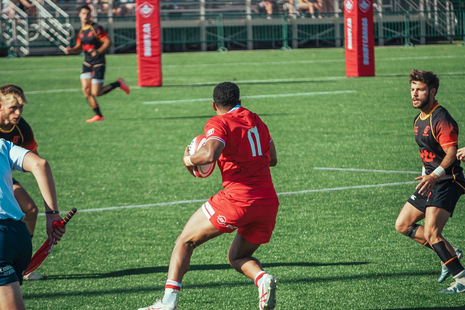 Canadian rugby players in red and dark uniforms compete on an artificial turf field during daytime match.