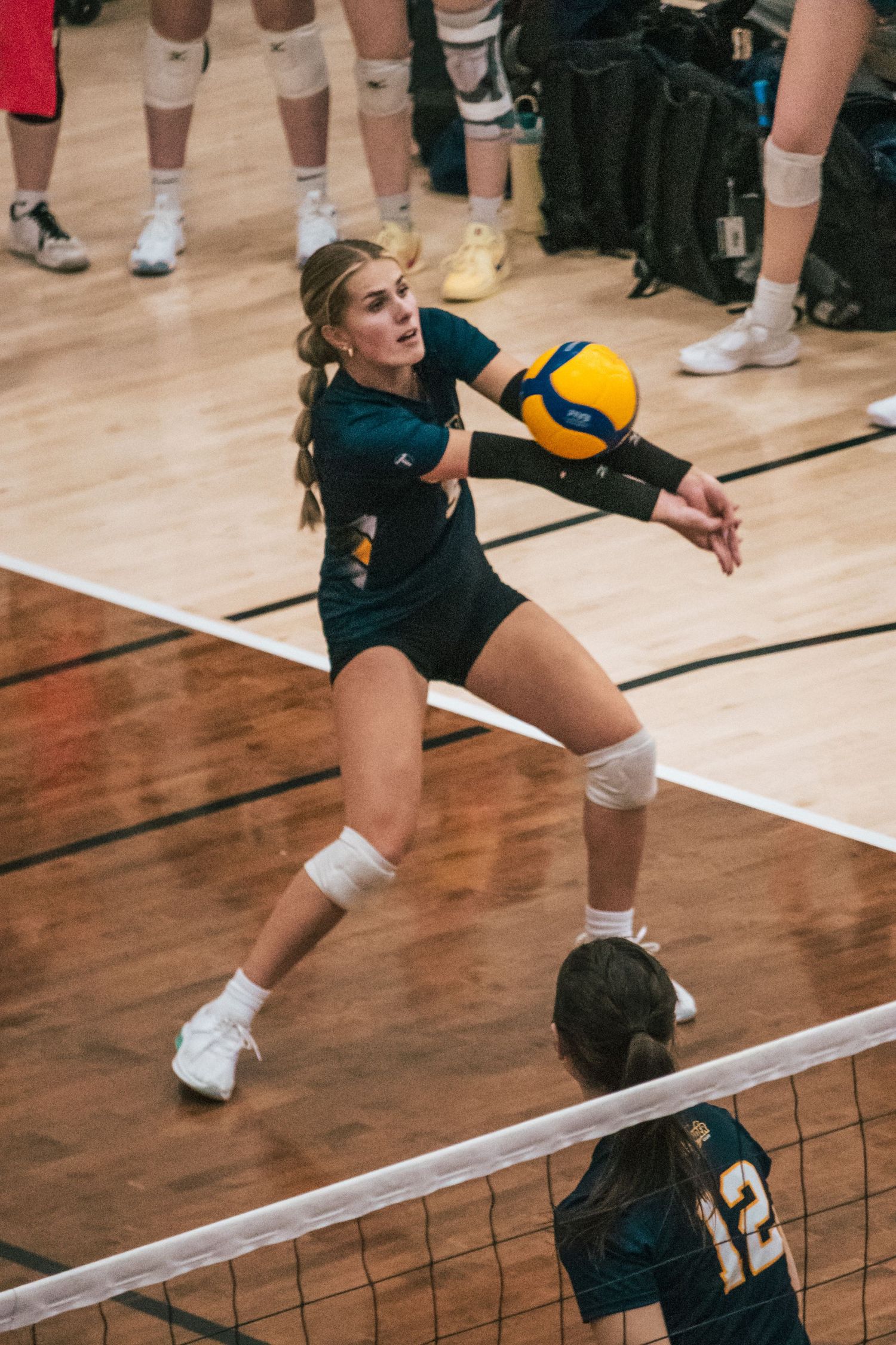 Volleyball player in dark uniform stretches to receive the ball during an indoor match on a wooden court.