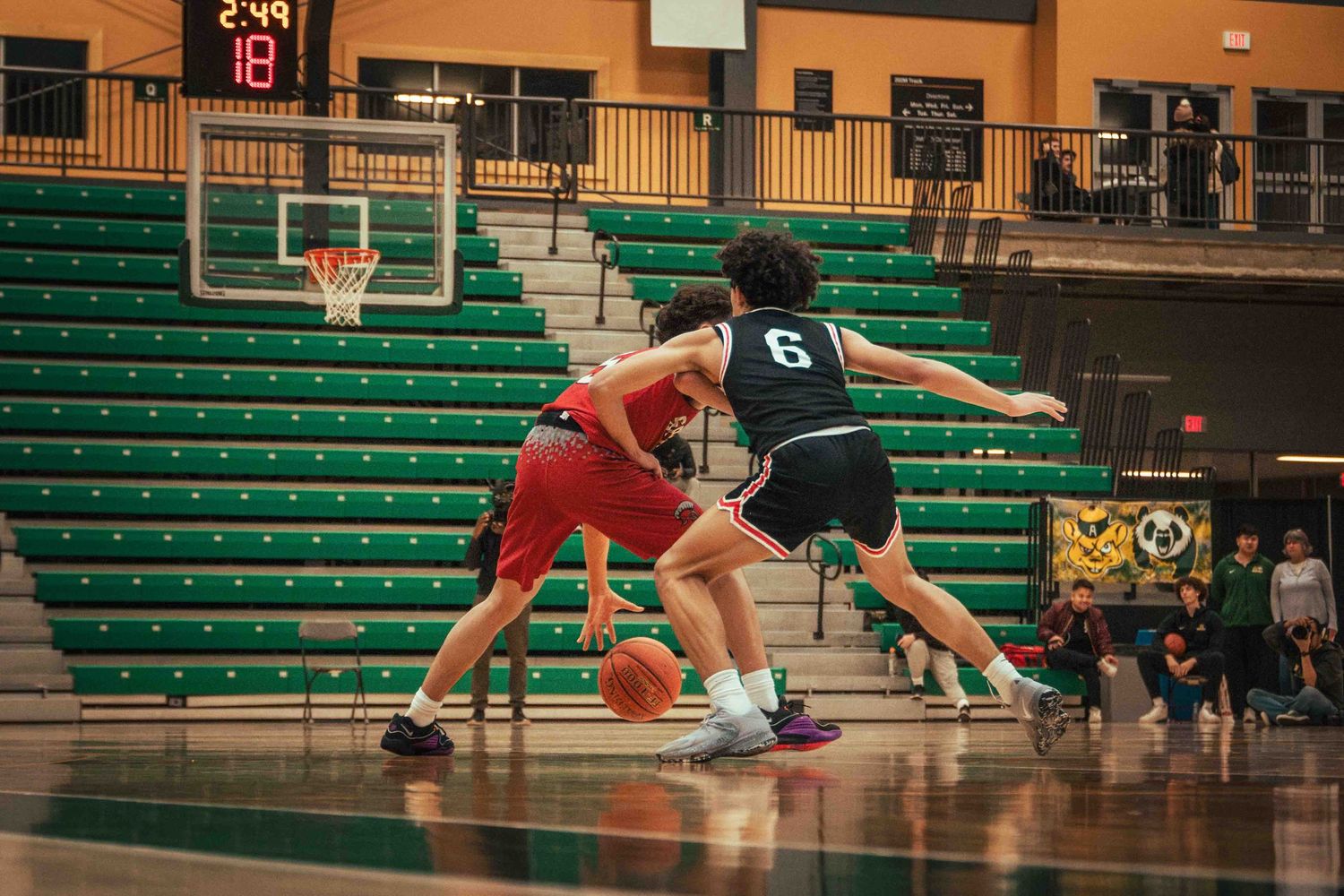 Basketball players compete in a gymnasium with green bleachers as one player dribbles past a defender.