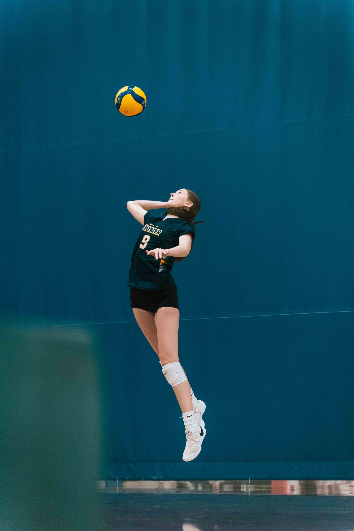 Volleyball player in black uniform jumps to serve against blue background.