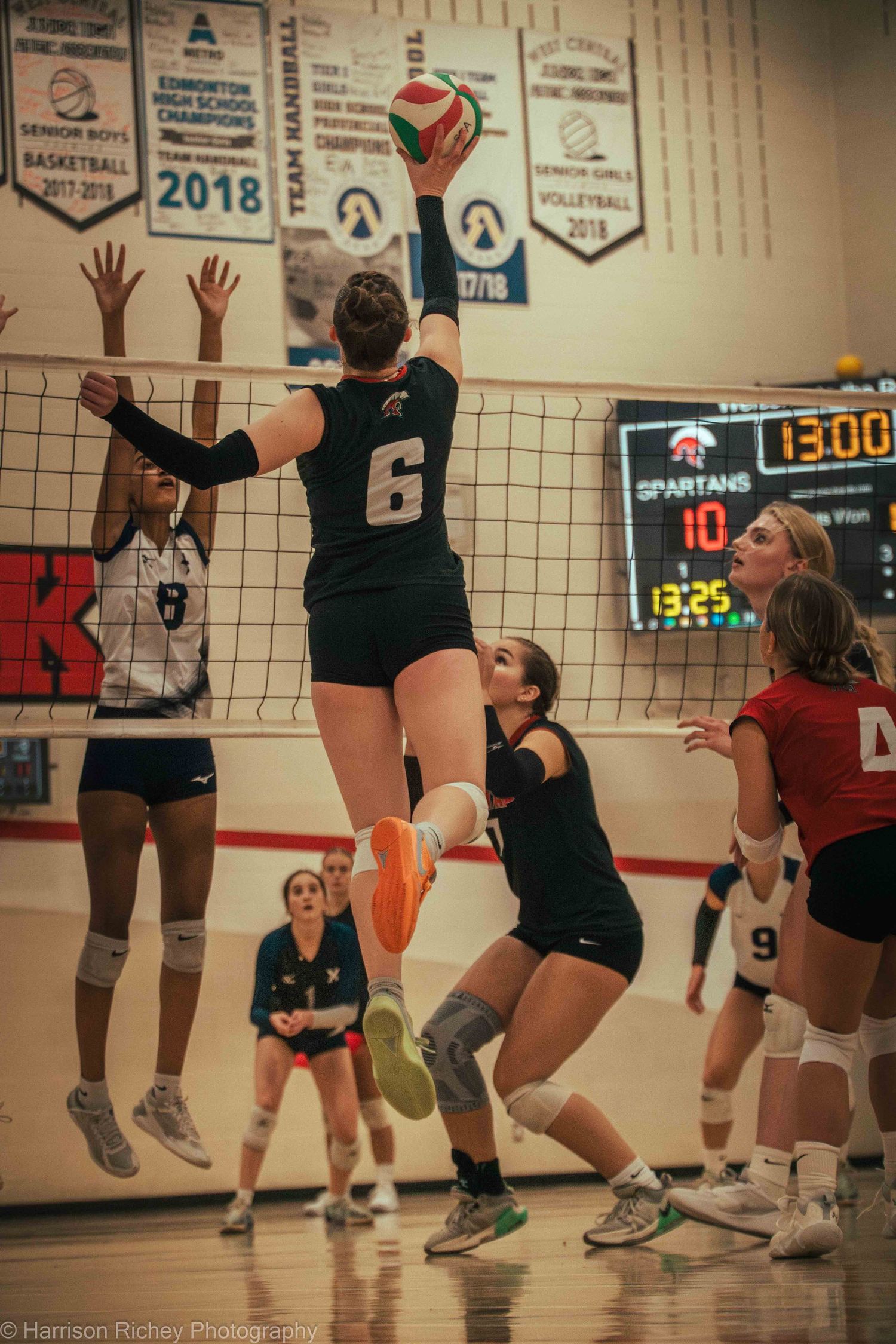 Players leap at the volleyball net during an indoor match with championship banners visible on the wall.