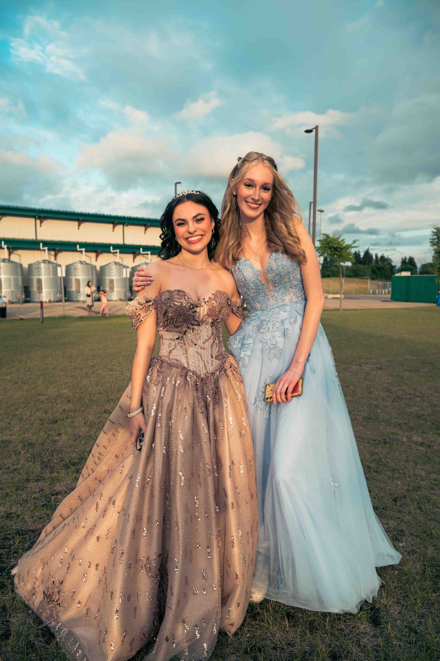 Two people in formal gowns, one sparkly brown and one light blue, pose together outdoors.