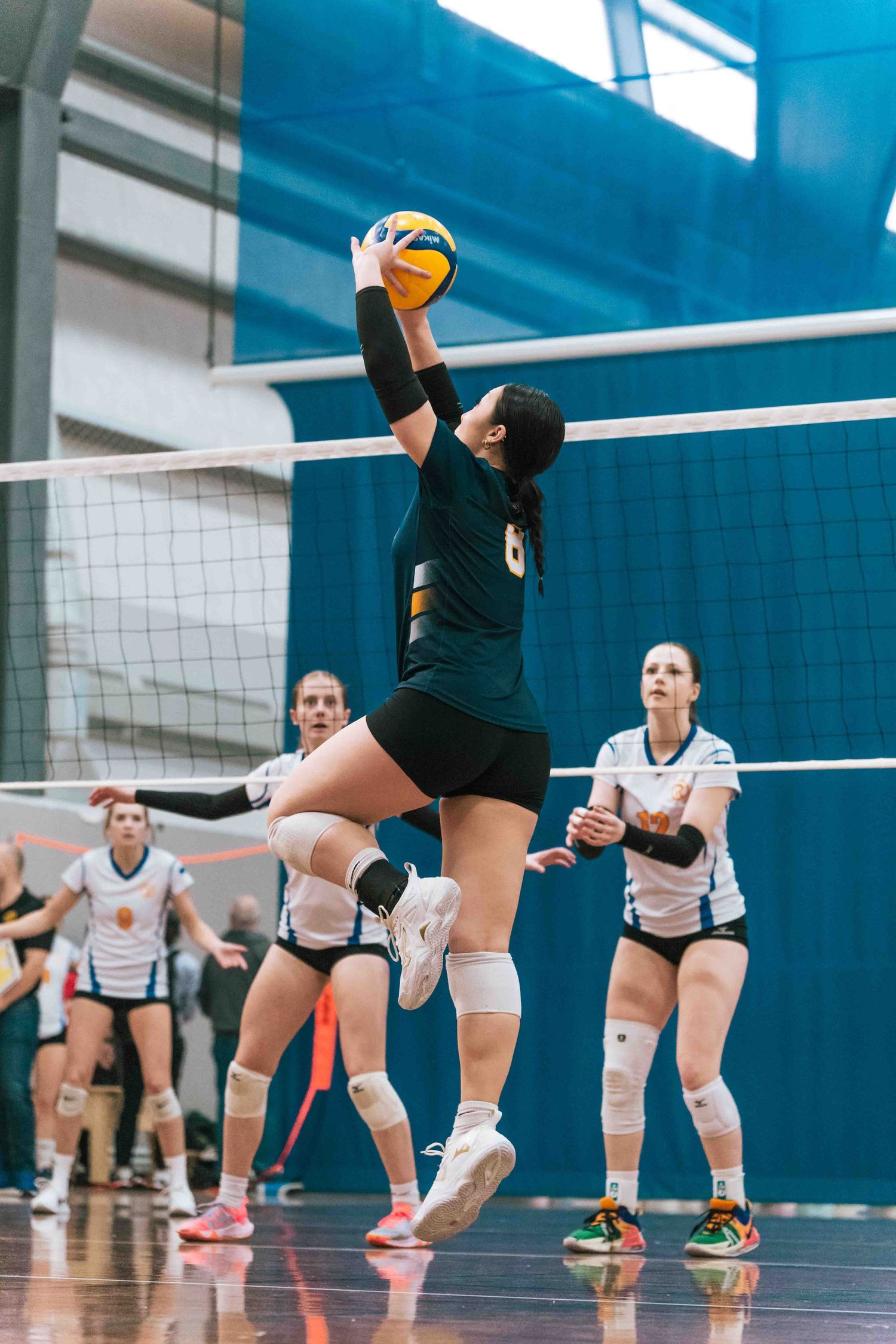 Volleyball player in dark jersey sets the ball during an indoor match against white-uniformed opponents.