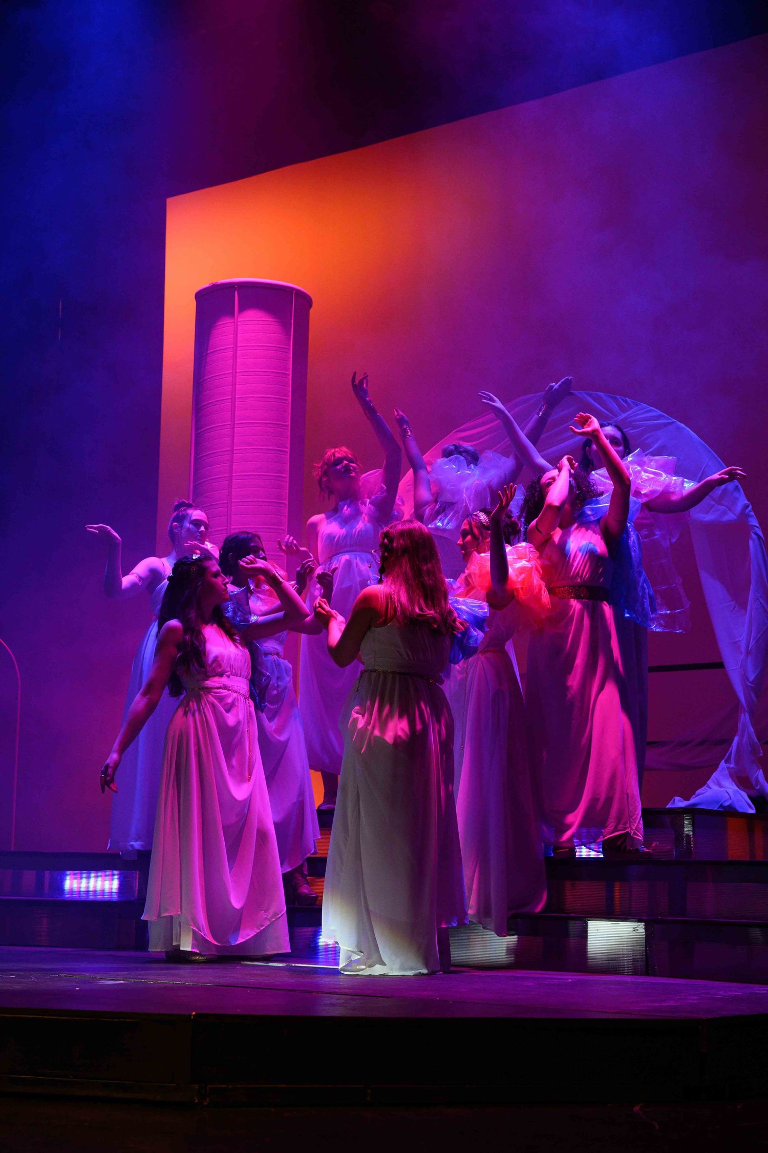 Dancers in flowing white dresses perform an expressive routine under dramatic purple and red stage lighting.