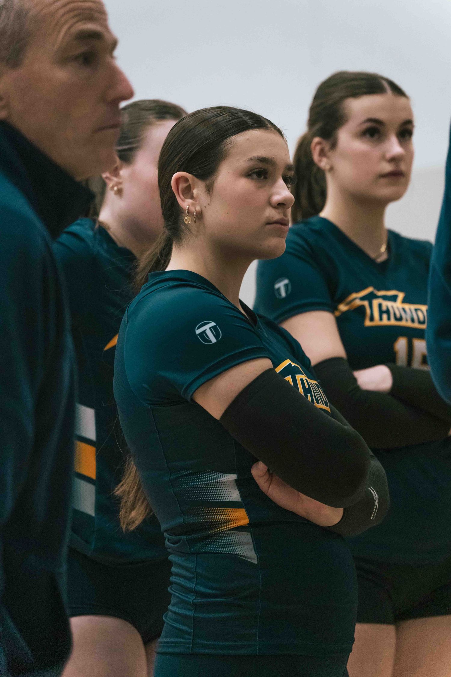 Team stands on sideline wearing dark green uniforms during volleyball match.
