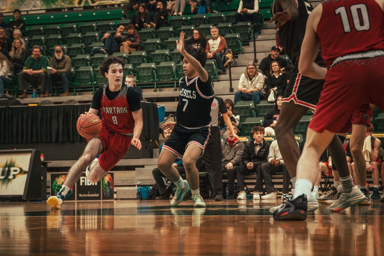Basketball players compete during an indoor game with one player in red dribbling past a defender in black.