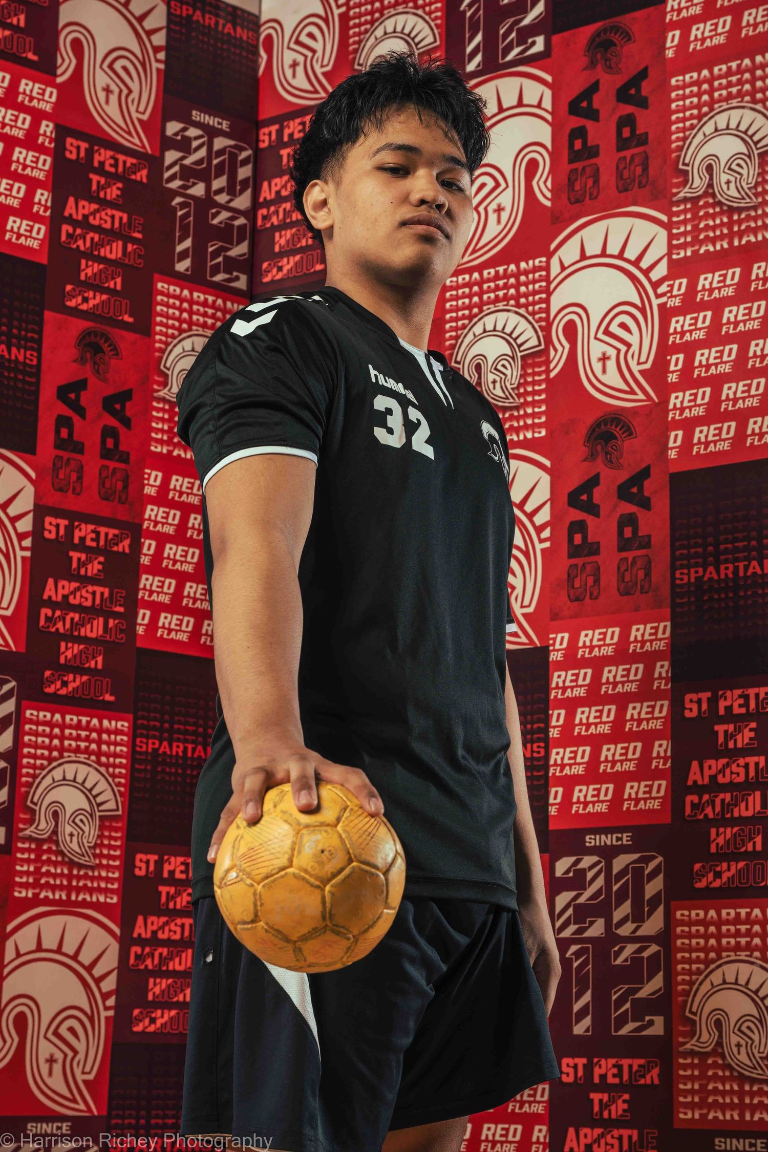 Handball player in black jersey poses with golden ball against red branded backdrop.