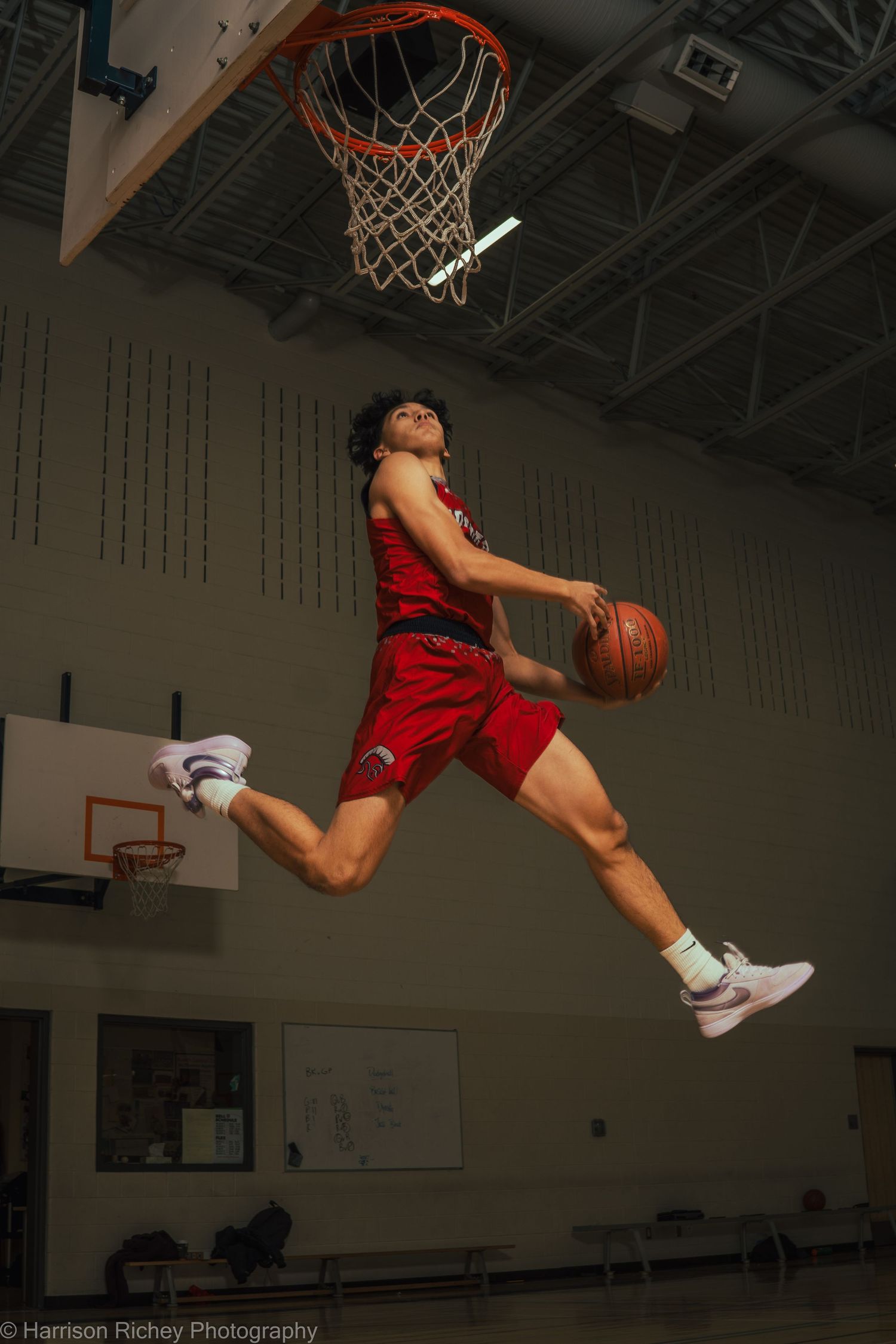 Basketball player in red uniform leaping high for a slam dunk in indoor gym.