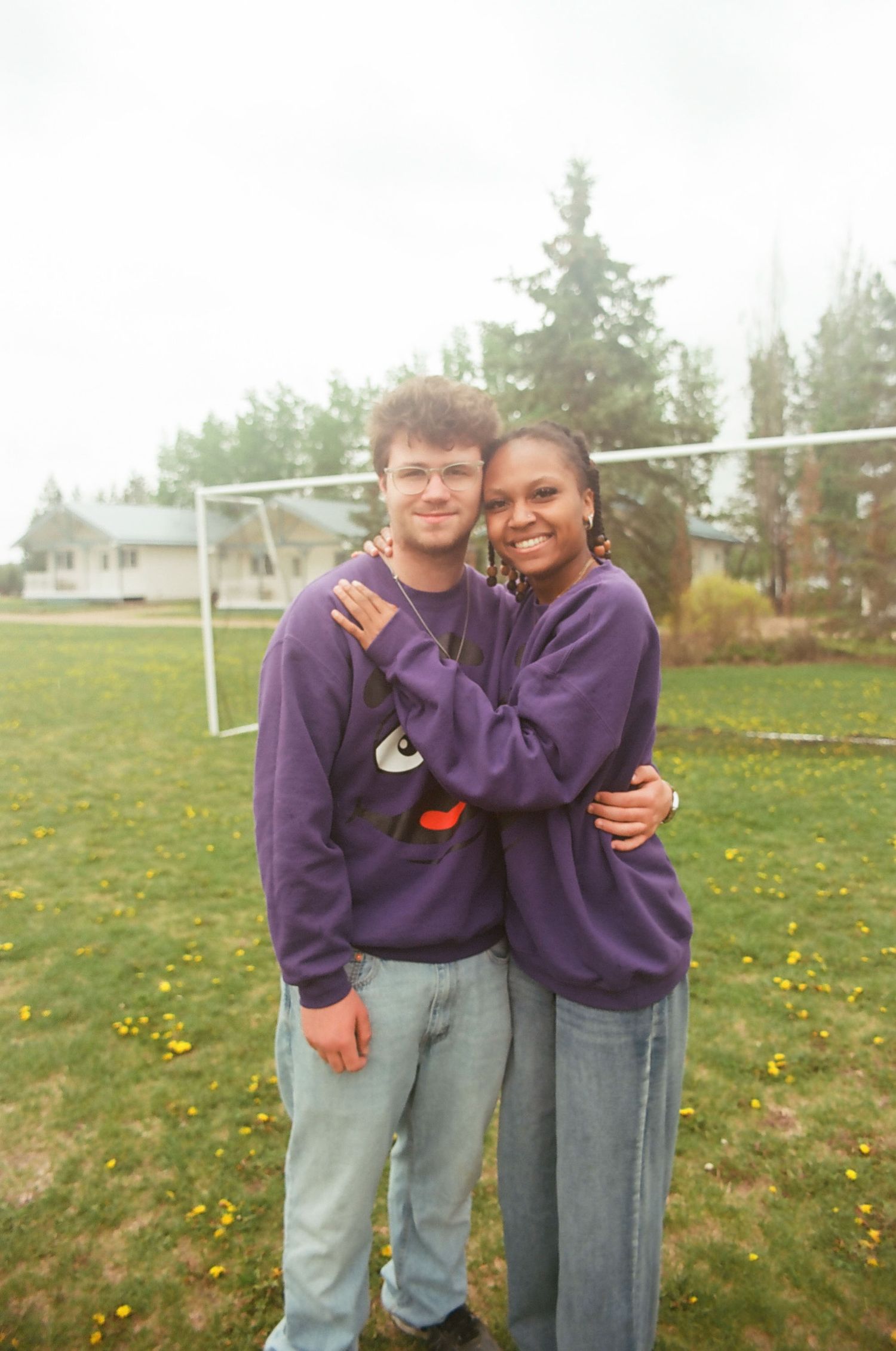 Two people in matching purple sweatshirts pose together on a grassy field with dandelions.