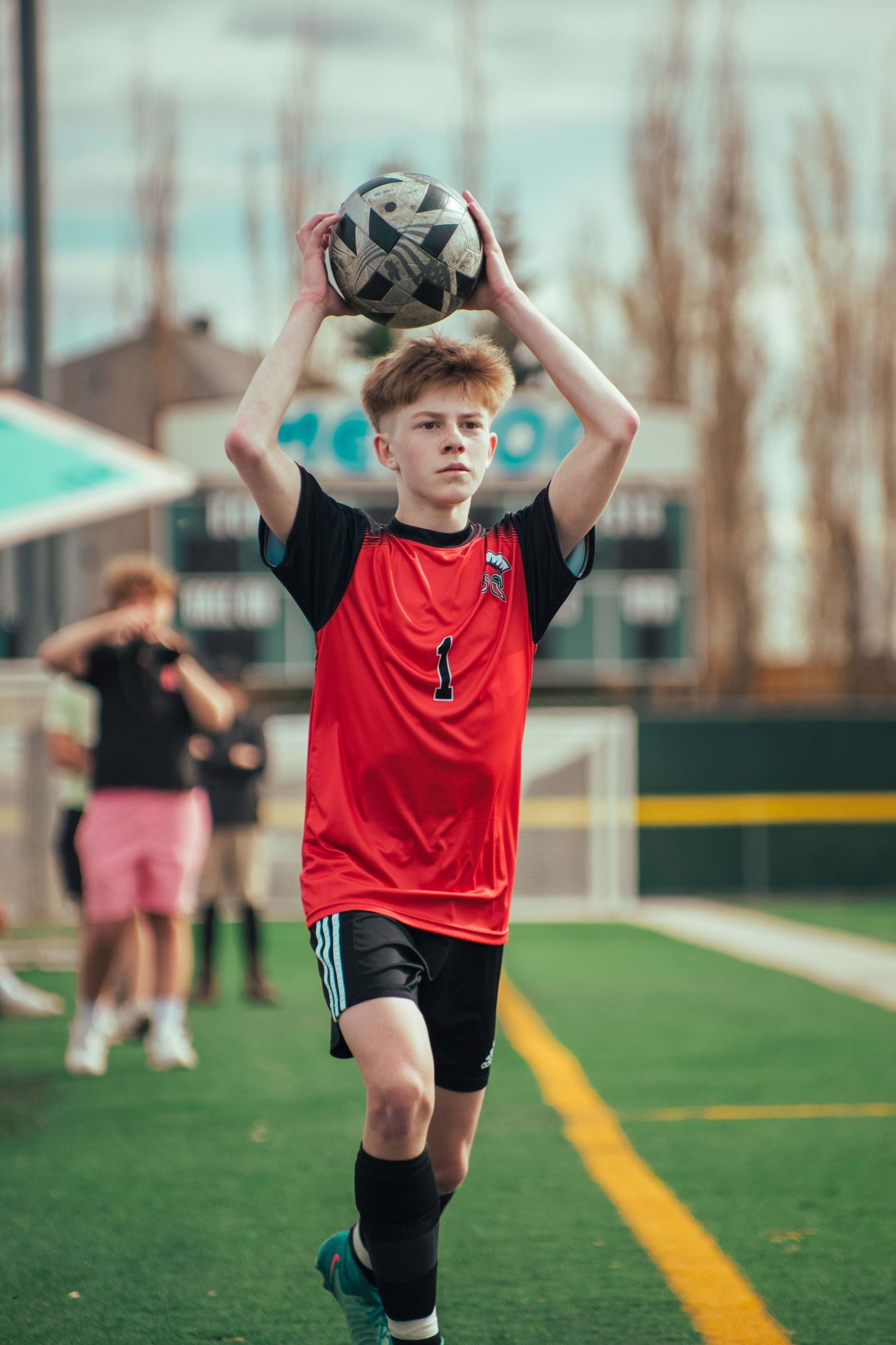 Young soccer player in red jersey preparing to throw in the ball on a sports field.