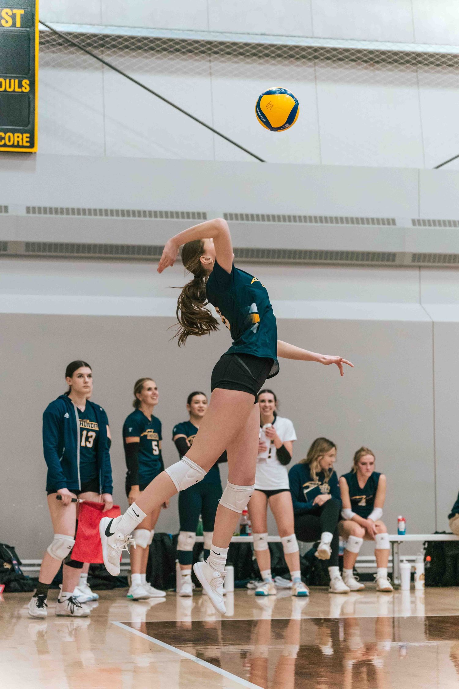 Volleyball player jumps high to strike ball during indoor game with teammates watching from sideline.