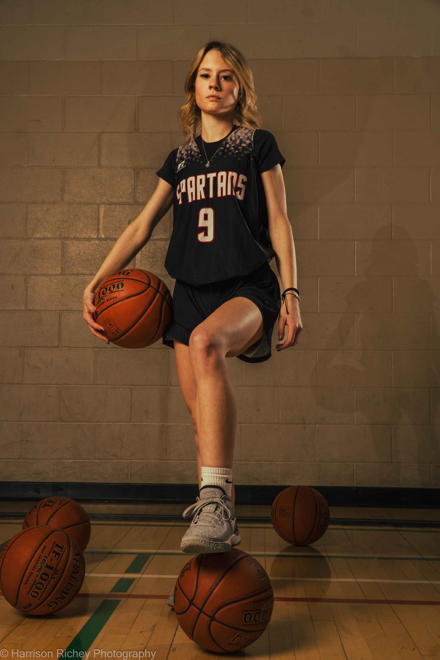 Basketball player sits on multiple basketballs on gym floor in dramatic lighting.