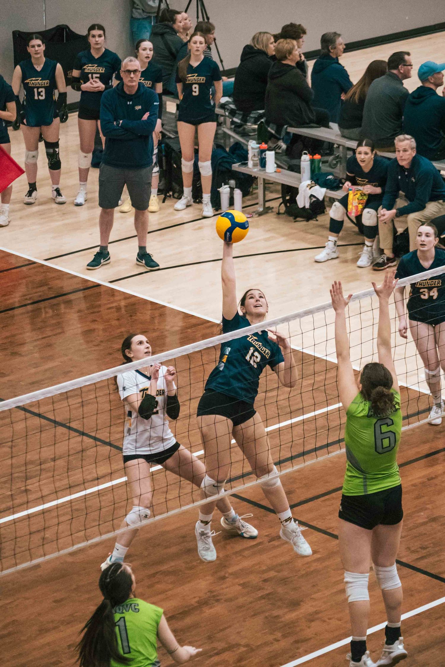 Volleyball players compete at the net during match with team benches visible in background.